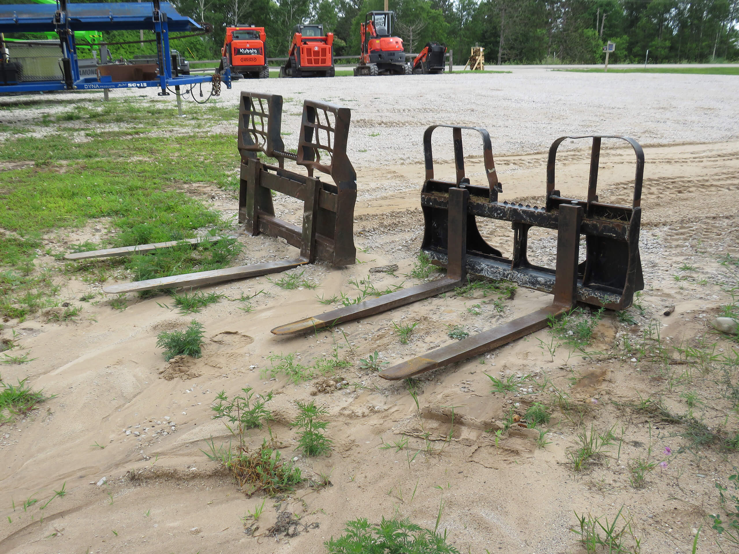 Two metal forklift attachments rest on sandy ground with parked construction vehicles in the background.