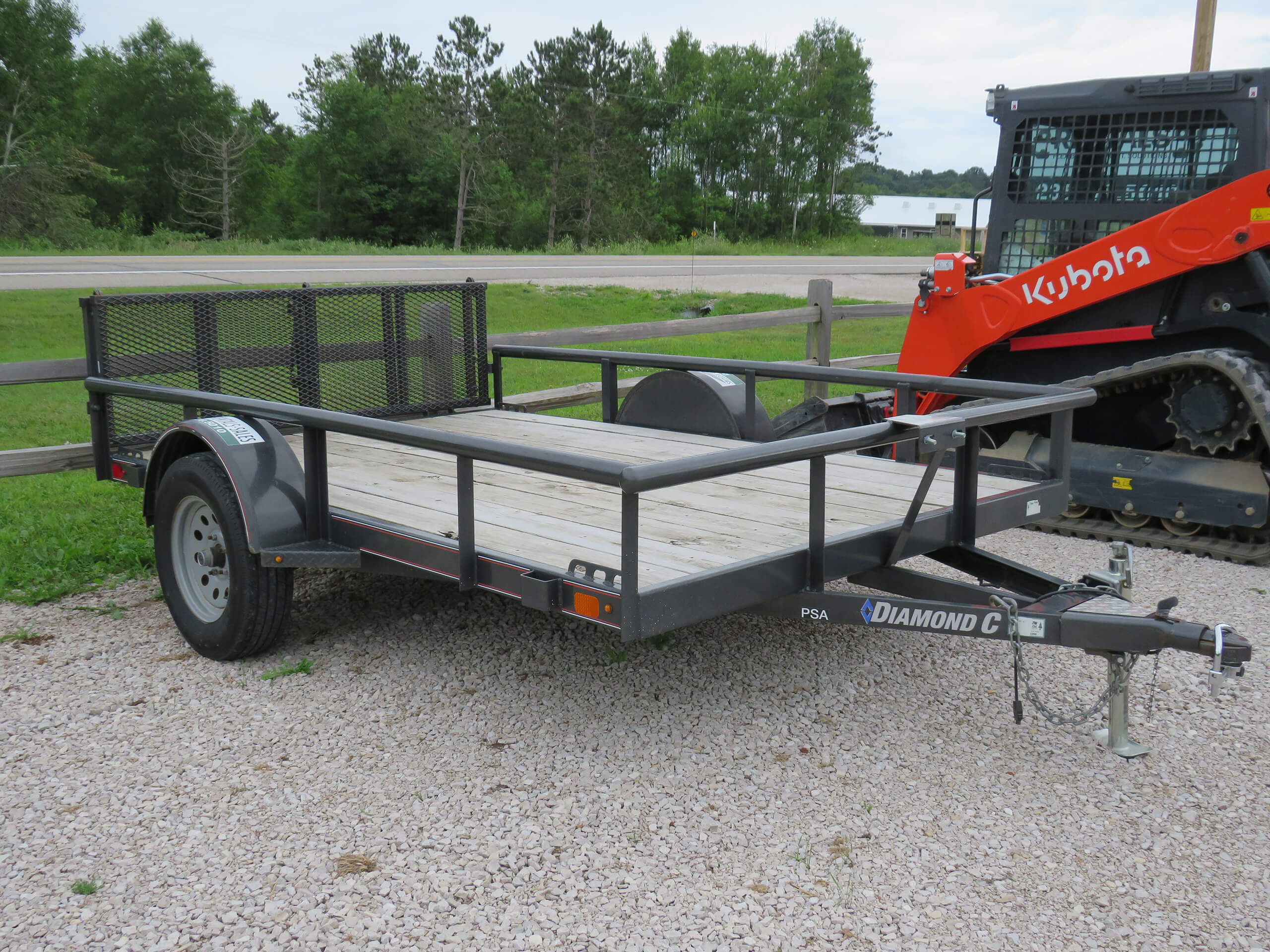 A flatbed utility trailer with mesh sides and a wooden floor is parked on gravel next to a fence, with construction equipment visible in the background.