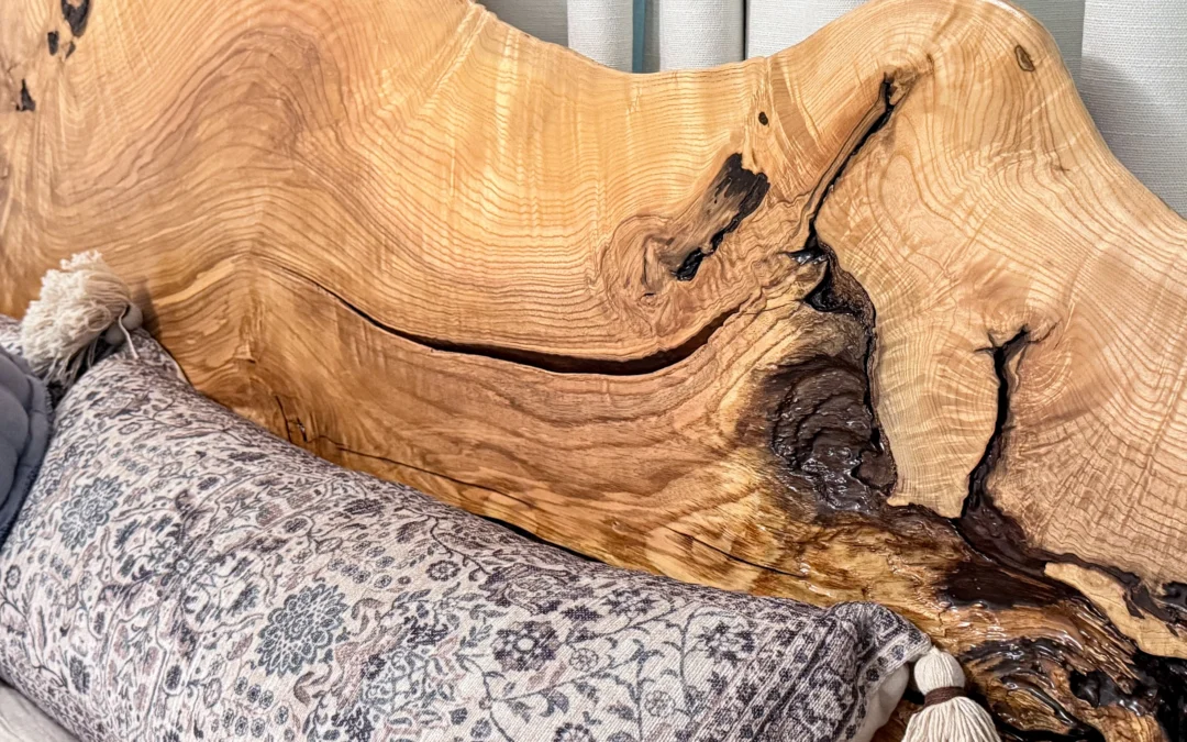Brookside Custom Woodcraft Fine Furniture & Custom Cabinetry Penn Yan NY A wooden headboard with a natural, irregular edge and visible grain patterns behind a bed with a patterned pillow and tassels. Light-colored curtains hang in the background.