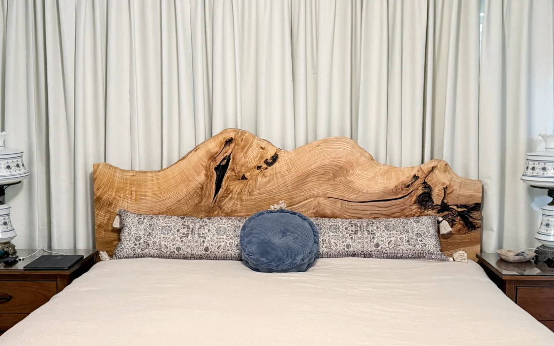 Brookside Custom Woodcraft Fine Furniture & Custom Cabinetry Penn Yan NY A neatly made bed with a live edge wooden headboard, two patterned pillows, a round blue pillow, and two nightstands with matching white lamps, set against closed cream curtains.