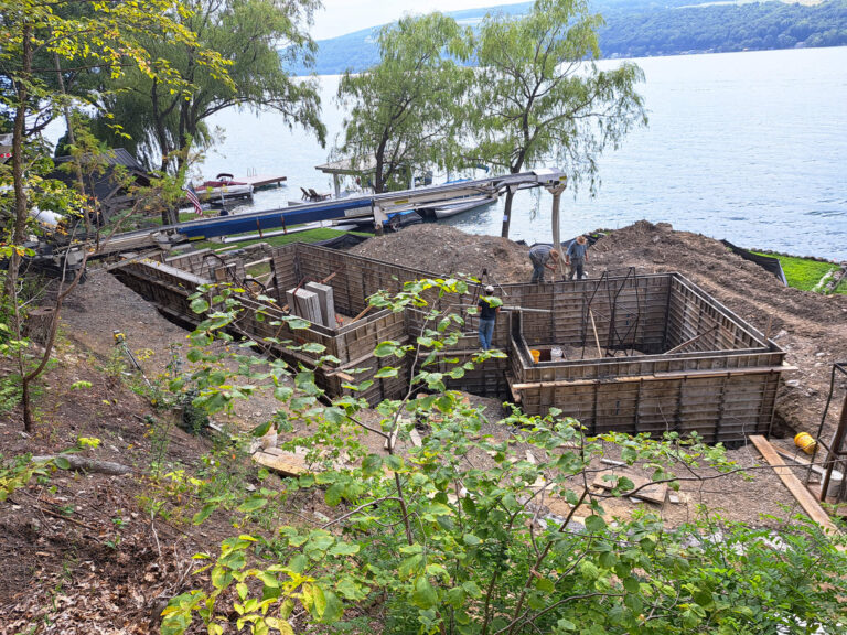 Eastview Poured Walls Concrete Flatwork Dundee, NY Construction site near a lakeshore with wooden formwork for concrete foundations; equipment and workers are present, surrounded by trees and dirt piles.