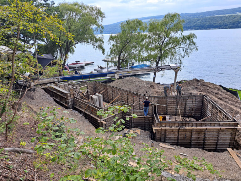 Eastview Poured Walls Concrete Flatwork Dundee, NY Workers build concrete forms for a foundation near a lake, with construction materials and machinery on a sloped, wooded shoreline.