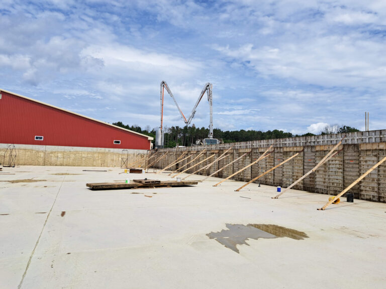 Eastview Poured Walls Concrete Flatwork Dundee, NY A construction site with a concrete foundation, wooden supports against a tall wall, scattered materials, and a red building to the left under a partly cloudy sky.