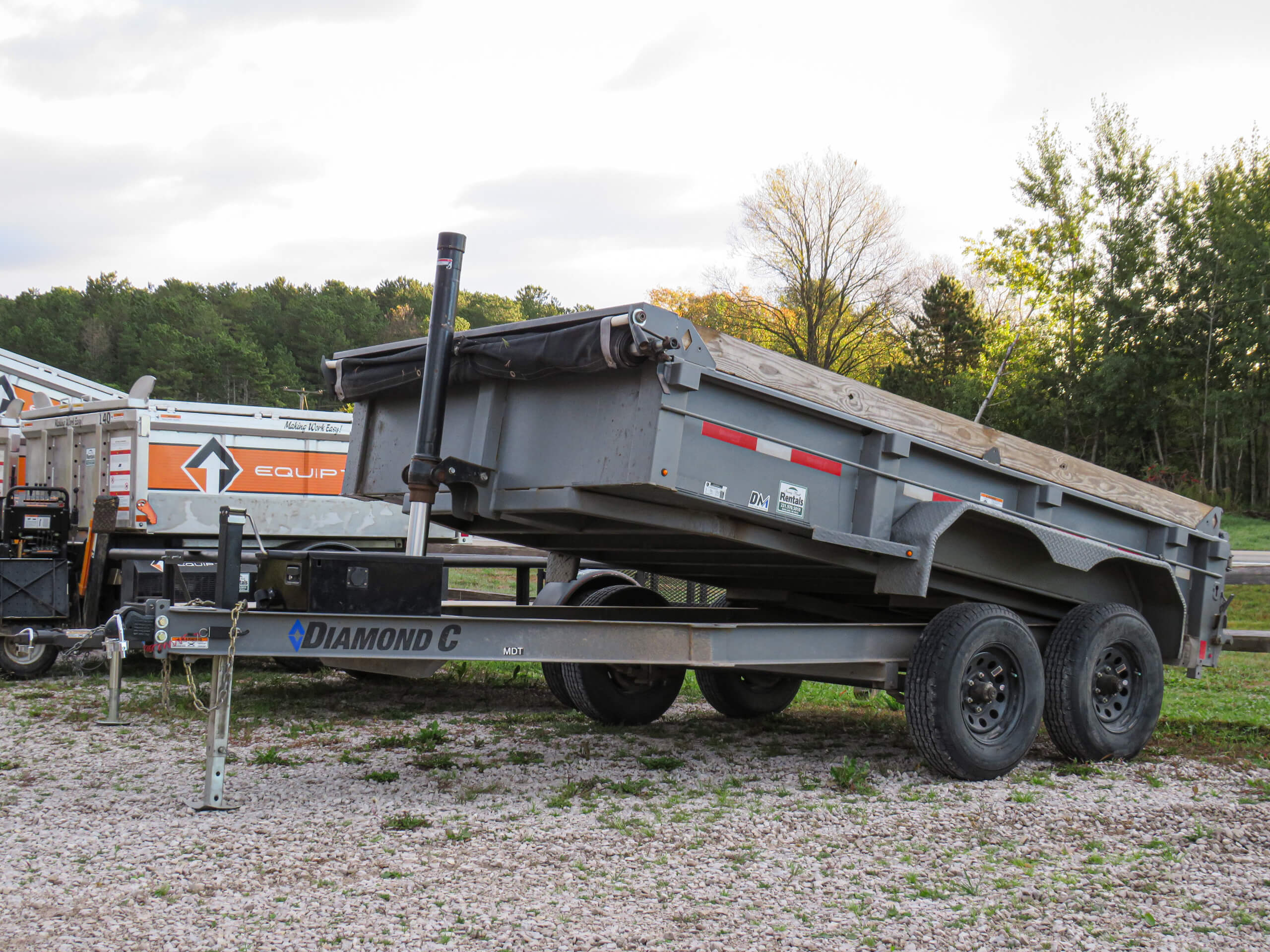A black Diamond C dump trailer with two axles is parked on gravel beside a white utility trailer and trees in the background.