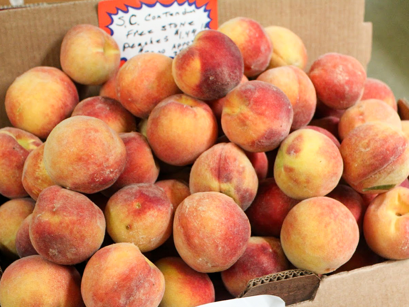 Country Variety Store Bellafontaine OH A pile of ripe peaches is displayed in a cardboard box, with a handwritten sign partially visible in the background.
