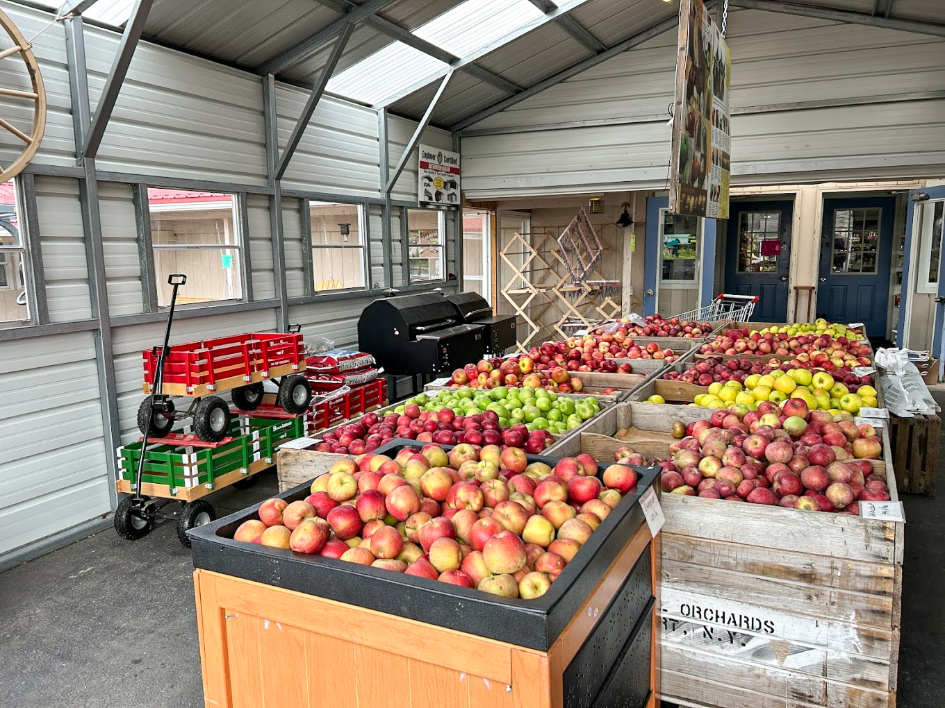 Country Variety Store Bellafontaine OH Interior of a farm market stand displaying bins of assorted apples, crates, a grill, a green wagon, and drying racks under a metal roof.
