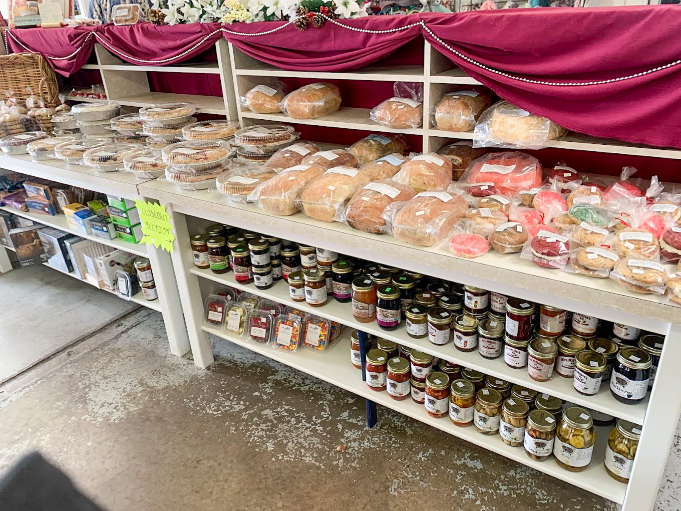 Country Variety Store Bellafontaine OH Shelves display assorted breads, pastries, and baked goods on top, with jars of jams, preserves, and other canned goods arranged on the lower shelves in a market setting.