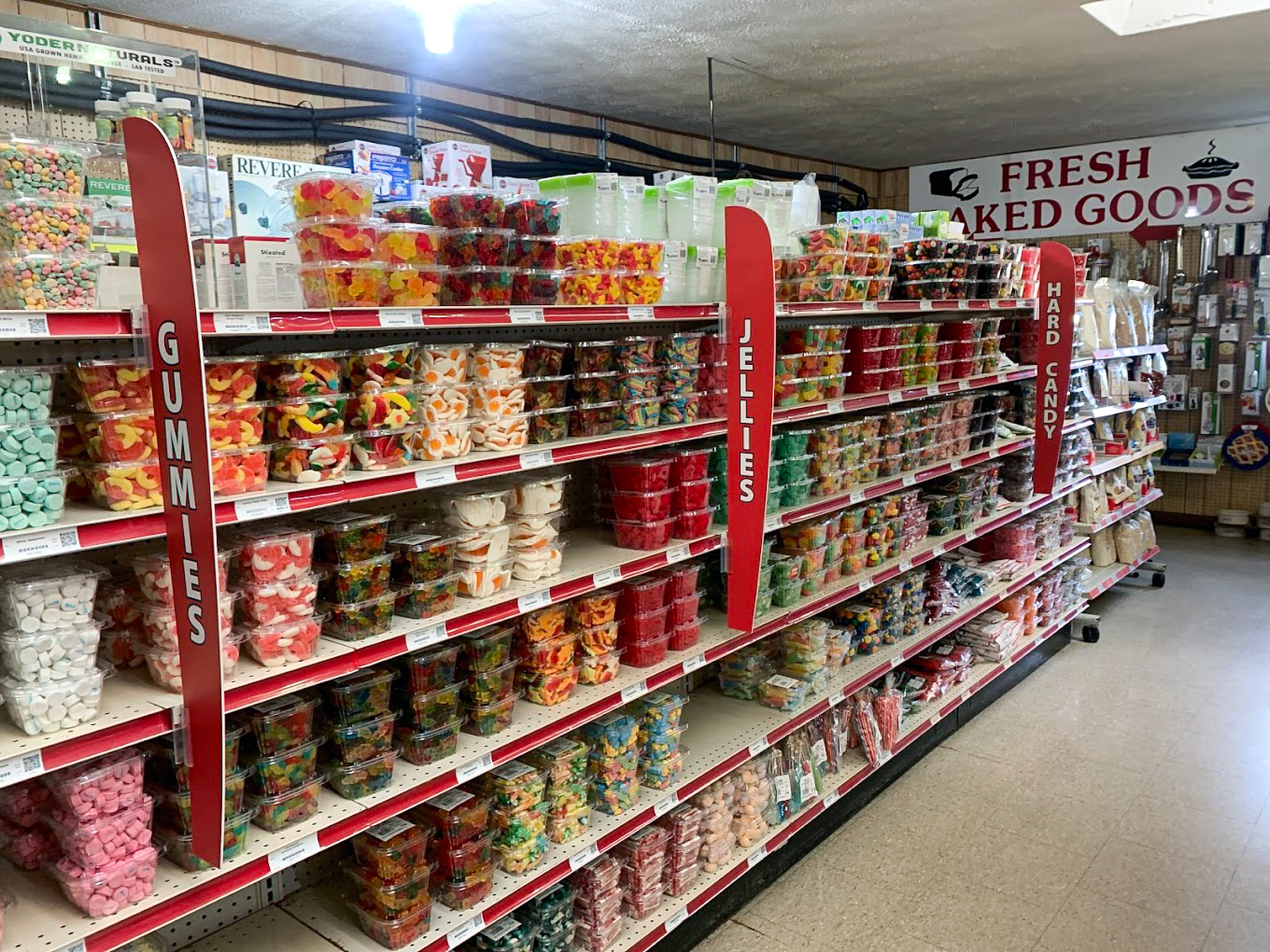 10.22.25 Country Variety Store Gallery 2-5 Store aisle with shelves filled with plastic containers of gummies, jellies, and hard candy; red signs label each candy section. A "Fresh Baked Goods" sign is visible in the background.