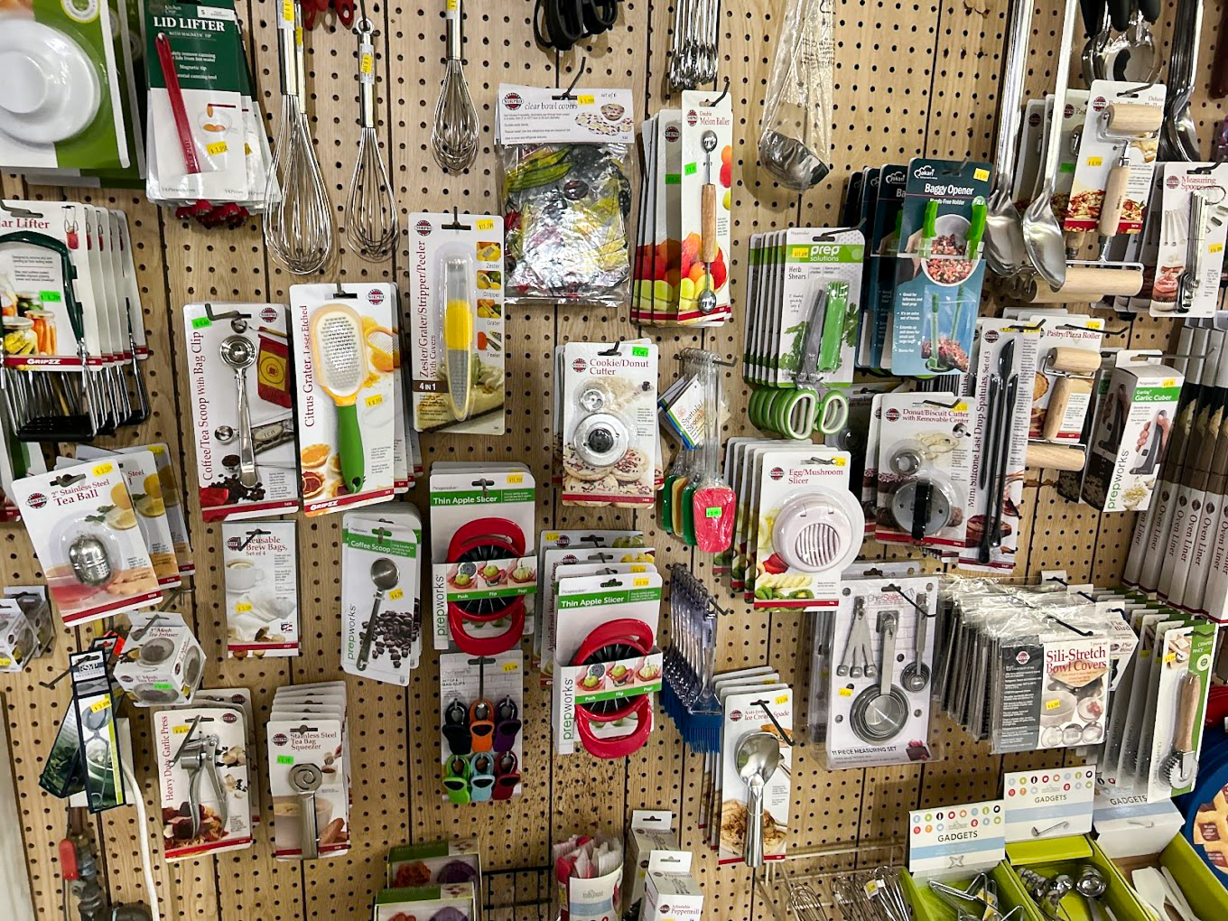 10.22.25 Country Variety Store Gallery 2-6 Assorted kitchen gadgets and utensils in packaging are displayed on metal hooks attached to a perforated pegboard wall in a store.
