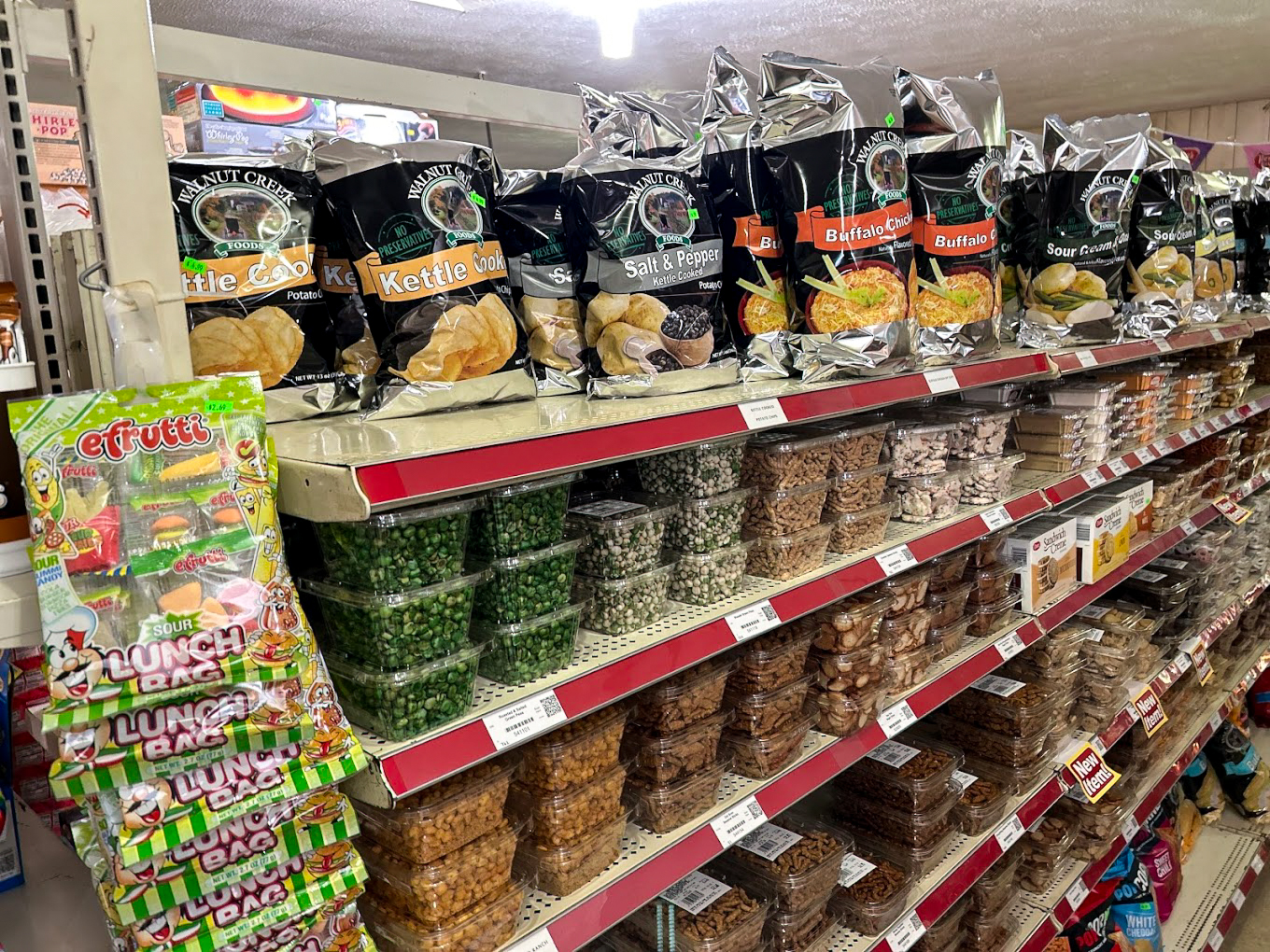 10.22.25 Country Variety Store Gallery 2-7 Shelves in a grocery store stocked with chips, nuts, snacks in plastic containers, and hanging packs of eFrutti gummy candy.