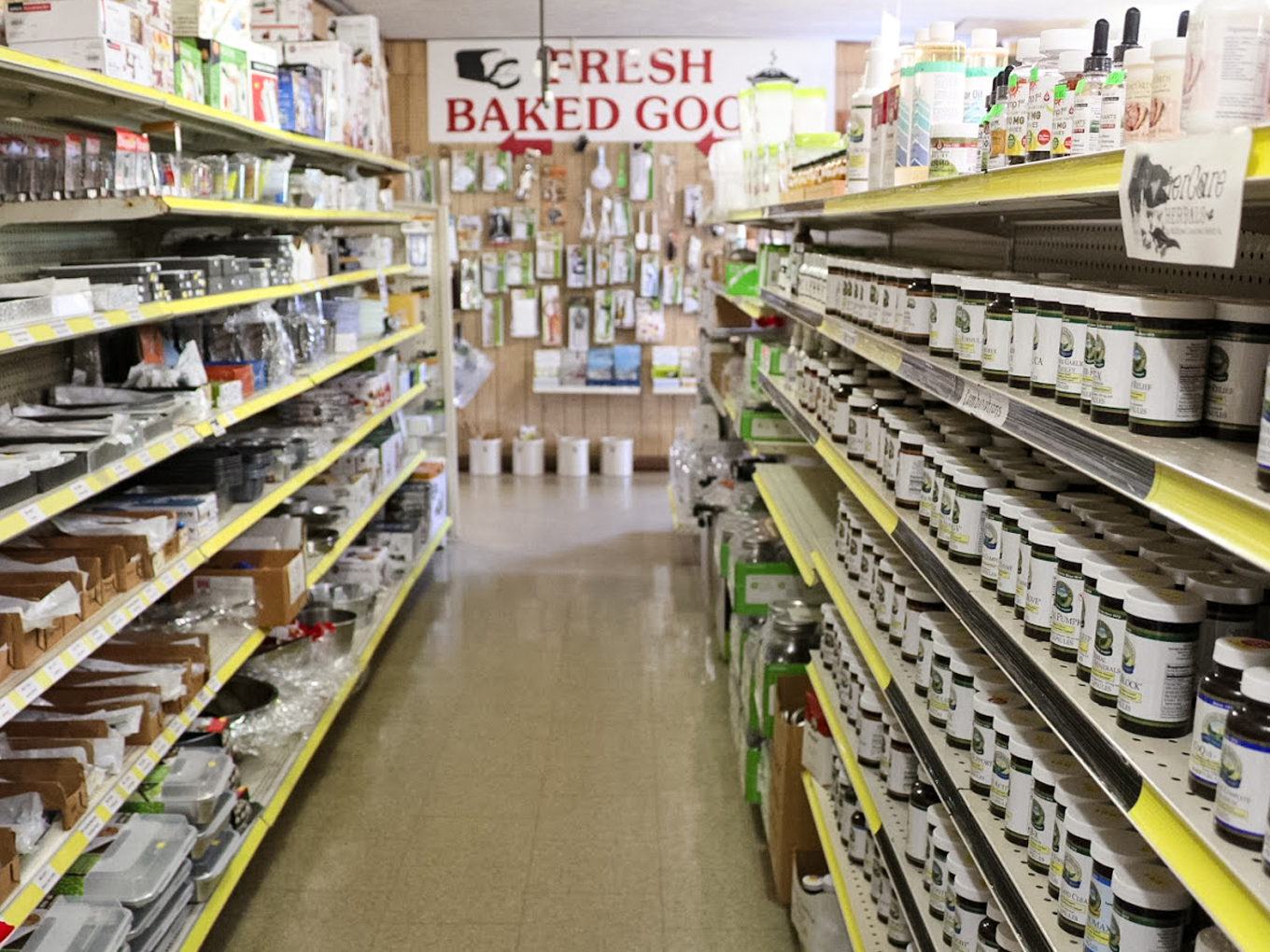 Country Variety Store Bellafontaine OH A grocery store aisle with shelves stocked with jars, containers, and kitchen supplies; a "Fresh Baked Goods" sign on the back wall.