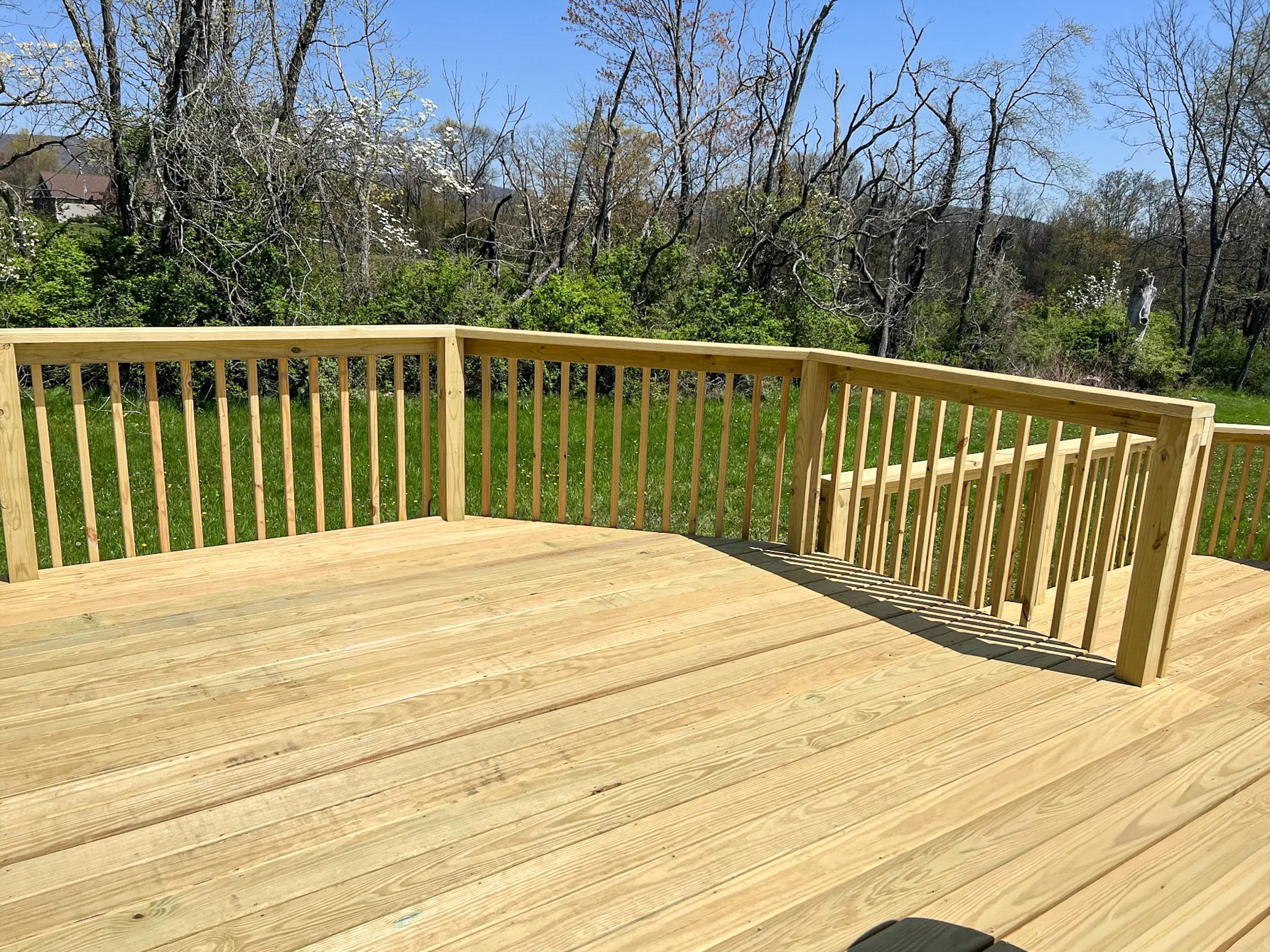 HMG Construction Mill Hall, PA A newly-built wooden deck with a railing and stairs overlooks a grassy yard with trees and shrubs under a clear sky.
