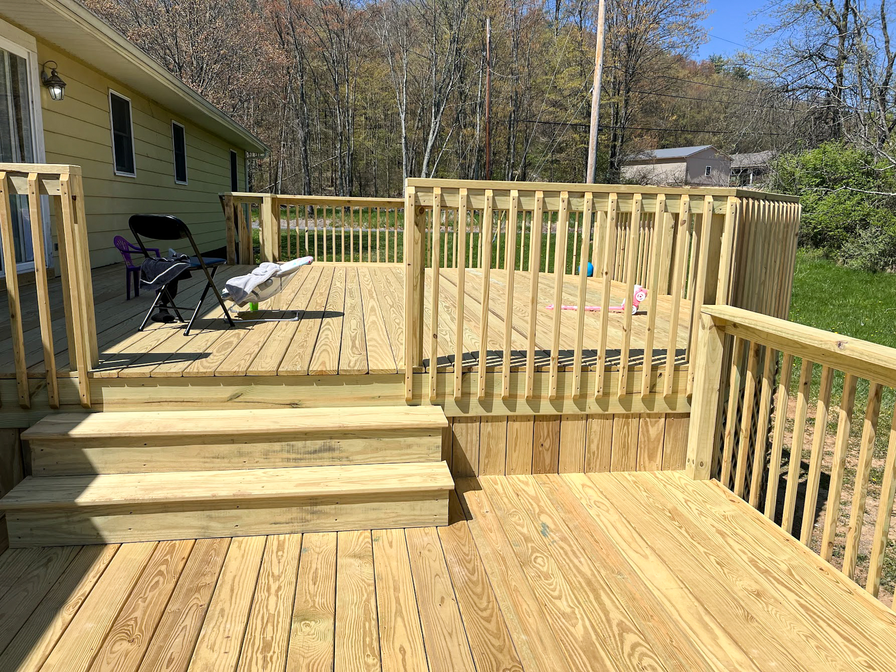 HMG Construction Mill Hall, PA A newly constructed wooden deck with steps, railings, and a foldable chair, attached to a yellow house, overlooking a grassy yard and trees.