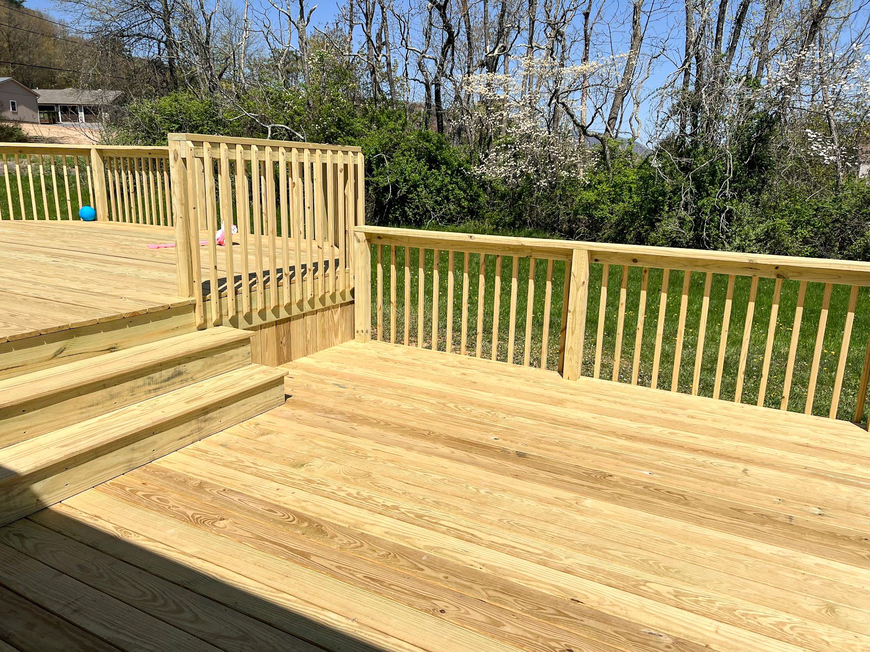 HMG Construction Mill Hall, PA A wooden deck with stairs and railings, overlooking grass and trees in the background under clear, sunny skies.