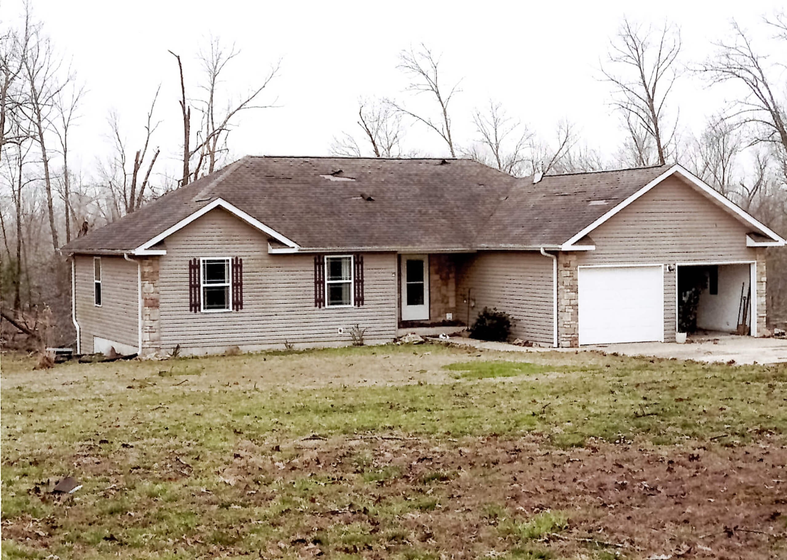 Single-story house with light-colored siding, attached two-car garage, pitched roof, and sparse trees in the background. The lawn is patchy with bare spots.