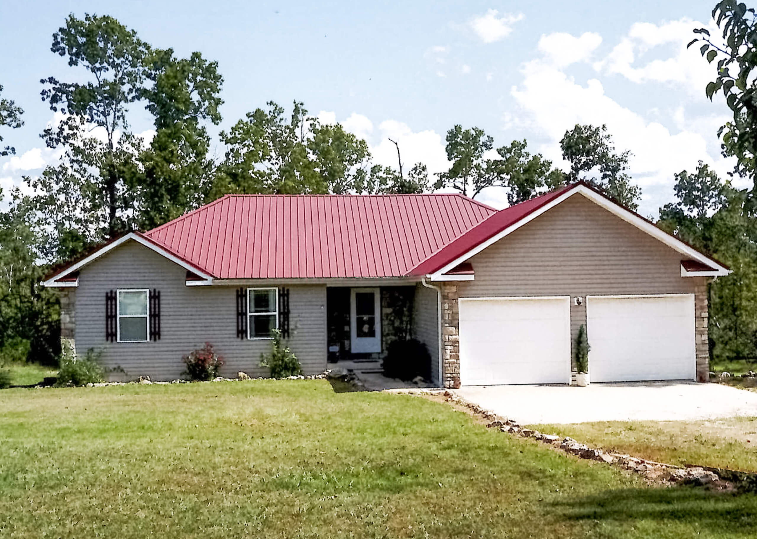 Single-story house with a red metal roof, beige siding, two-car garage, and a grassy front yard, surrounded by trees and a partly cloudy sky.