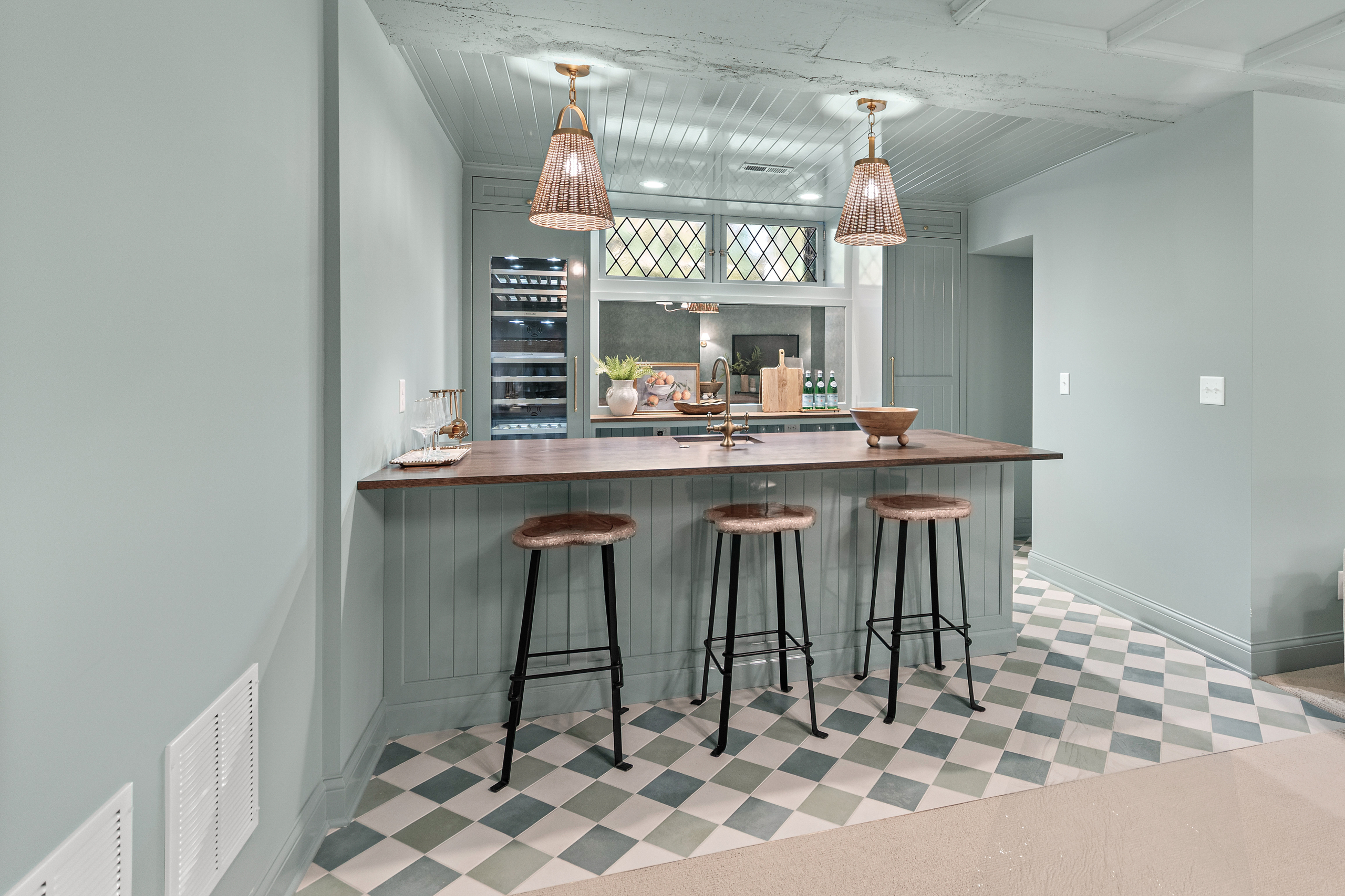 Kenrose Kitchen Kabinets Cabinetry Arthur IL A modern home bar with a wooden countertop, three bar stools, pendant lights, and checkered tile flooring in a soft green and white color scheme.