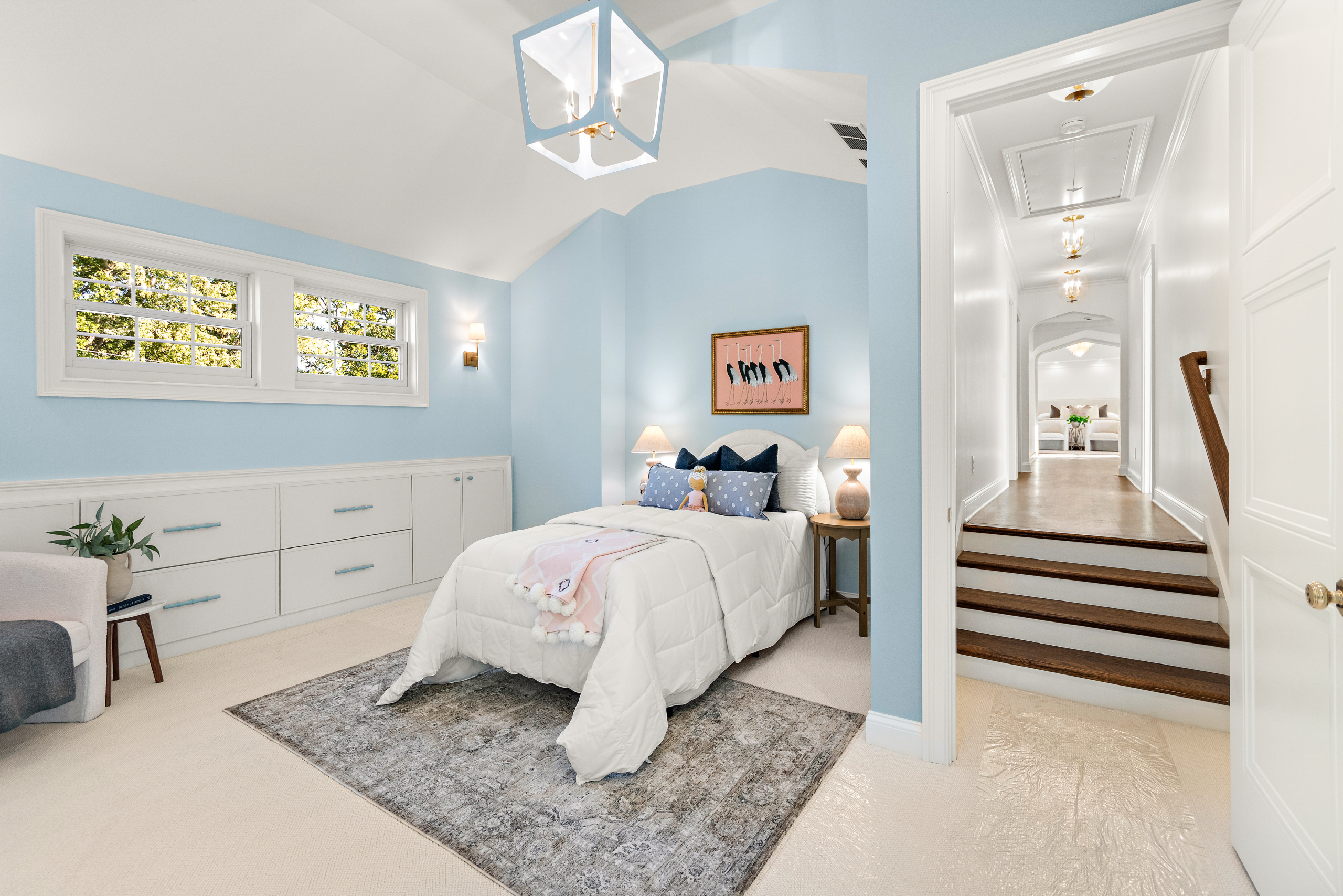 Kenrose Kitchen Kabinets Cabinetry Arthur IL A light blue bedroom with a bed, nightstands, and wall art; built-in drawers line one wall, and a hallway with steps leads to another room.