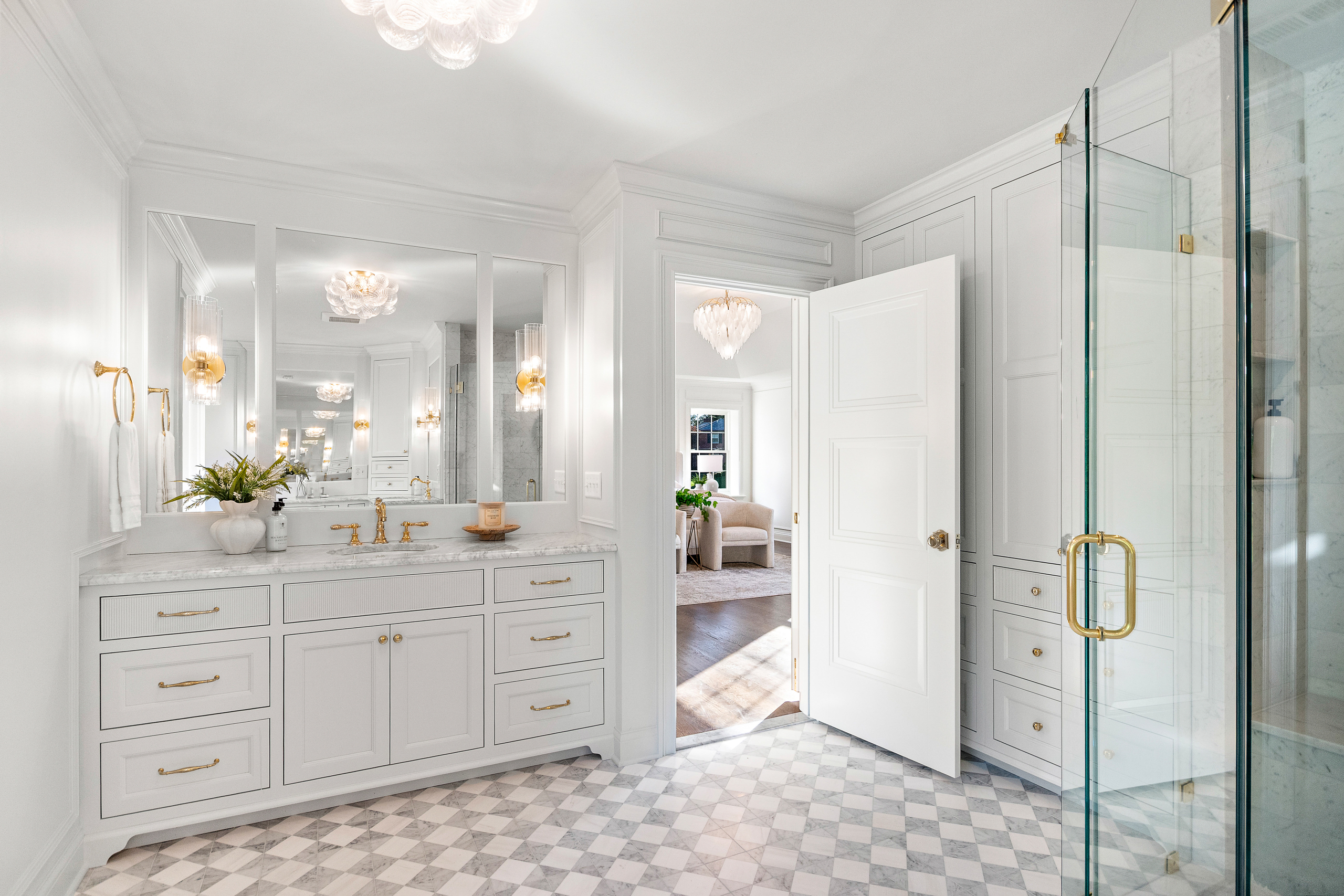 Kenrose Kitchen Kabinets Cabinetry Arthur IL Bright, modern bathroom with white cabinets, gold fixtures, marble countertop, large mirrors, glass shower, checkered tile floor, and an open door leading to a sunlit room.