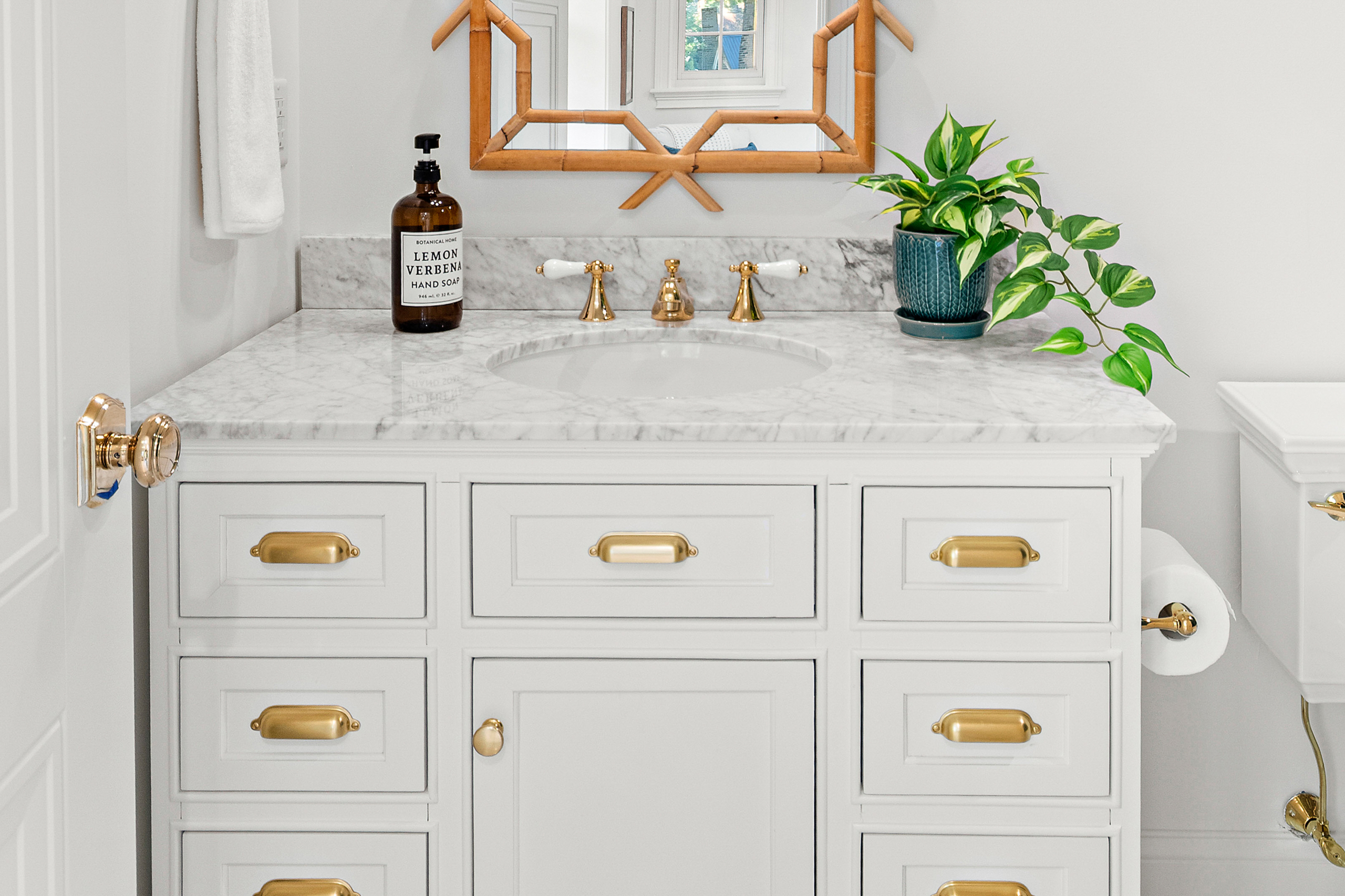Kenrose Kitchen Kabinets Cabinetry Arthur IL White bathroom vanity with gold hardware, marble countertop, undermount sink, soap dispenser, potted plant, towel, wall mirror, and toilet paper holder.