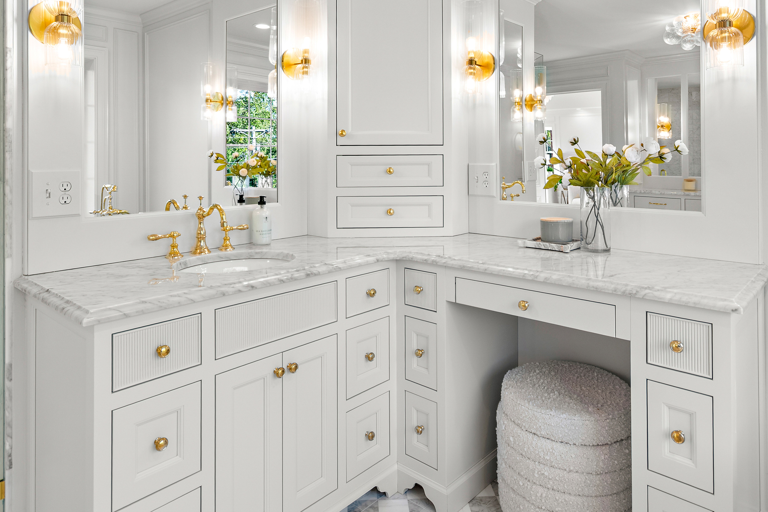 Kenrose Kitchen Kabinets Cabinetry Arthur IL Elegant white bathroom vanity with gold hardware, marble countertop, built-in sink, makeup area, flowers in vases, lit candle, and wall mirrors with gold sconces.