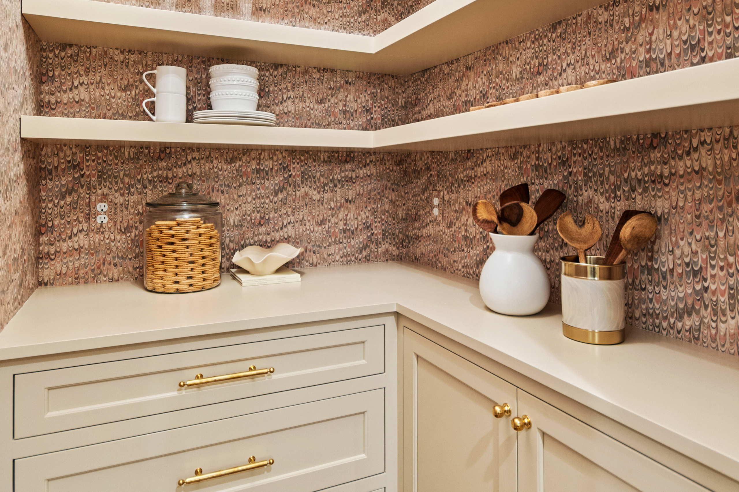 Kenrose Kitchen Kabinets Cabinetry Arthur IL Corner of a kitchen with cream cabinets, open shelves holding dishes, a glass jar of crackers, and containers with wooden utensils against a patterned backsplash.