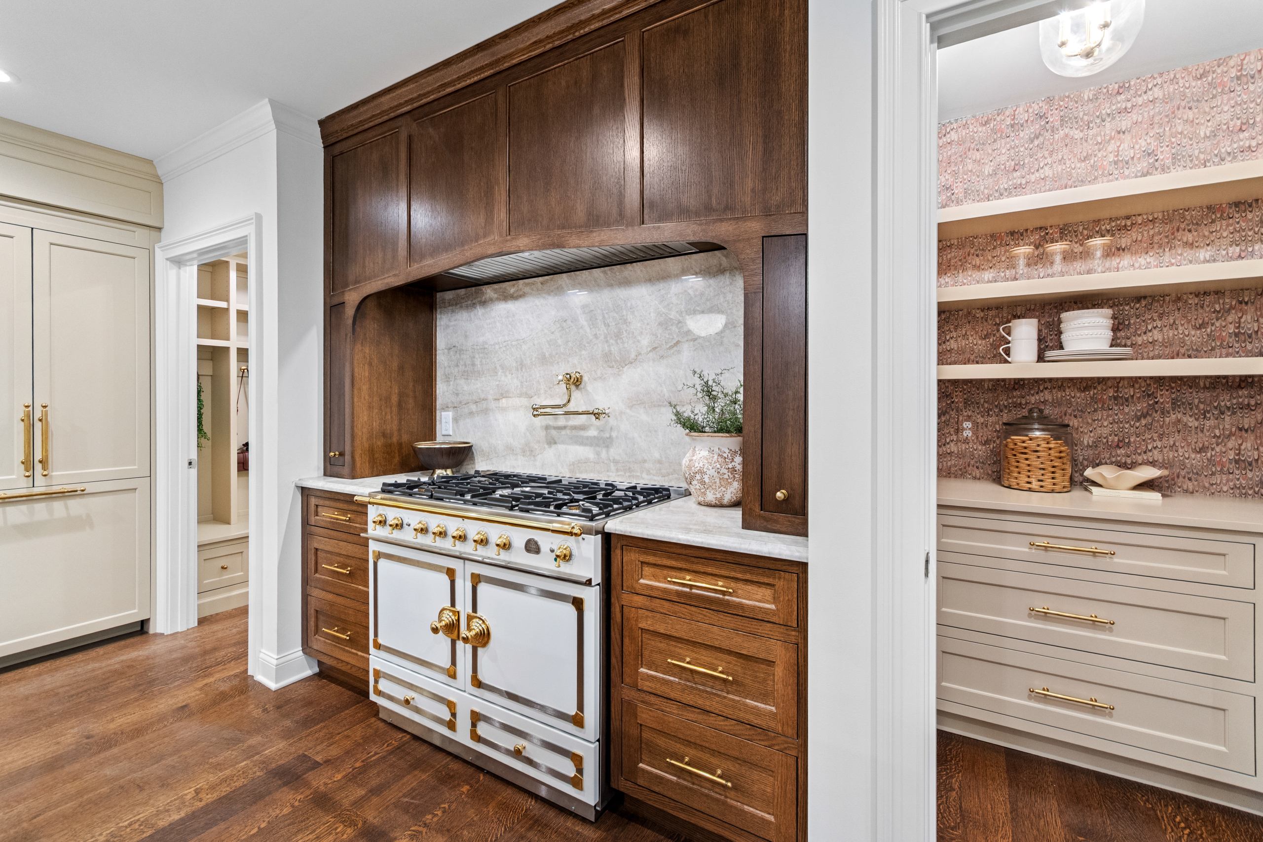 Kenrose Kitchen Kabinets Cabinetry Arthur IL A kitchen with dark wood cabinets, a white and gold range stove, marble backsplash, and an adjacent pantry with shelves holding dishes and decor items.