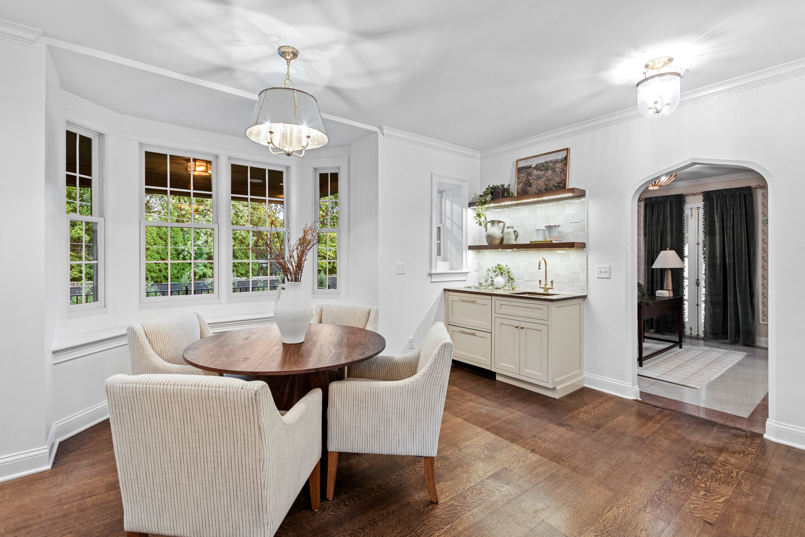Kenrose Kitchen Kabinets Cabinetry Arthur IL A dining area with a round wooden table, four upholstered chairs, a white vase centerpiece, large windows, hardwood floors, and a built-in sideboard with shelves.