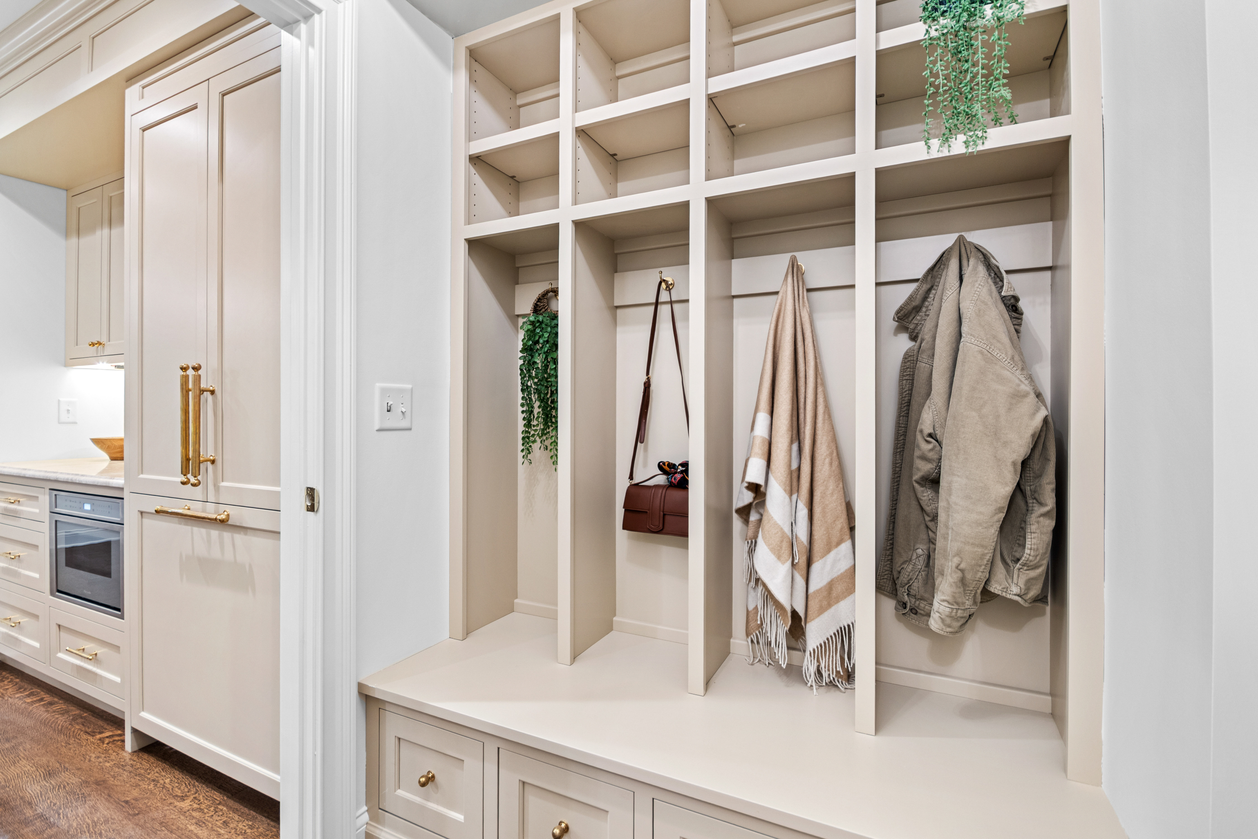 Kenrose Kitchen Kabinets Cabinetry Arthur IL Beige mudroom with built-in cubbies, hooks holding a jacket, scarf, and handbag, plus overhead shelves with potted plants. Kitchen is visible through adjacent doorway.