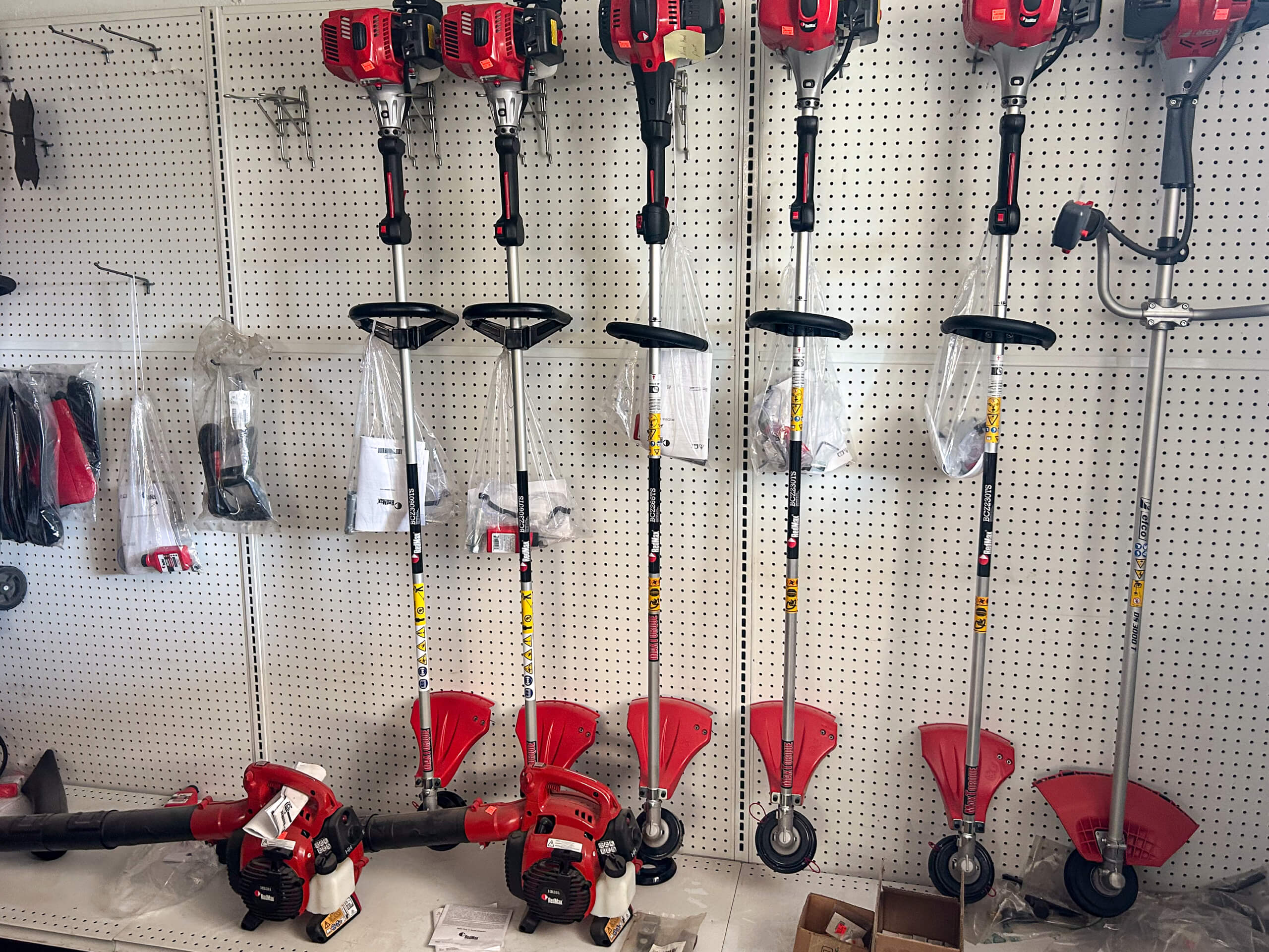 Buel Road Small Engine Repair Canajoharie, NY Red and silver string trimmers are displayed vertically and horizontally on a white pegboard wall in a store, with some accessories and parts on the lower shelf.