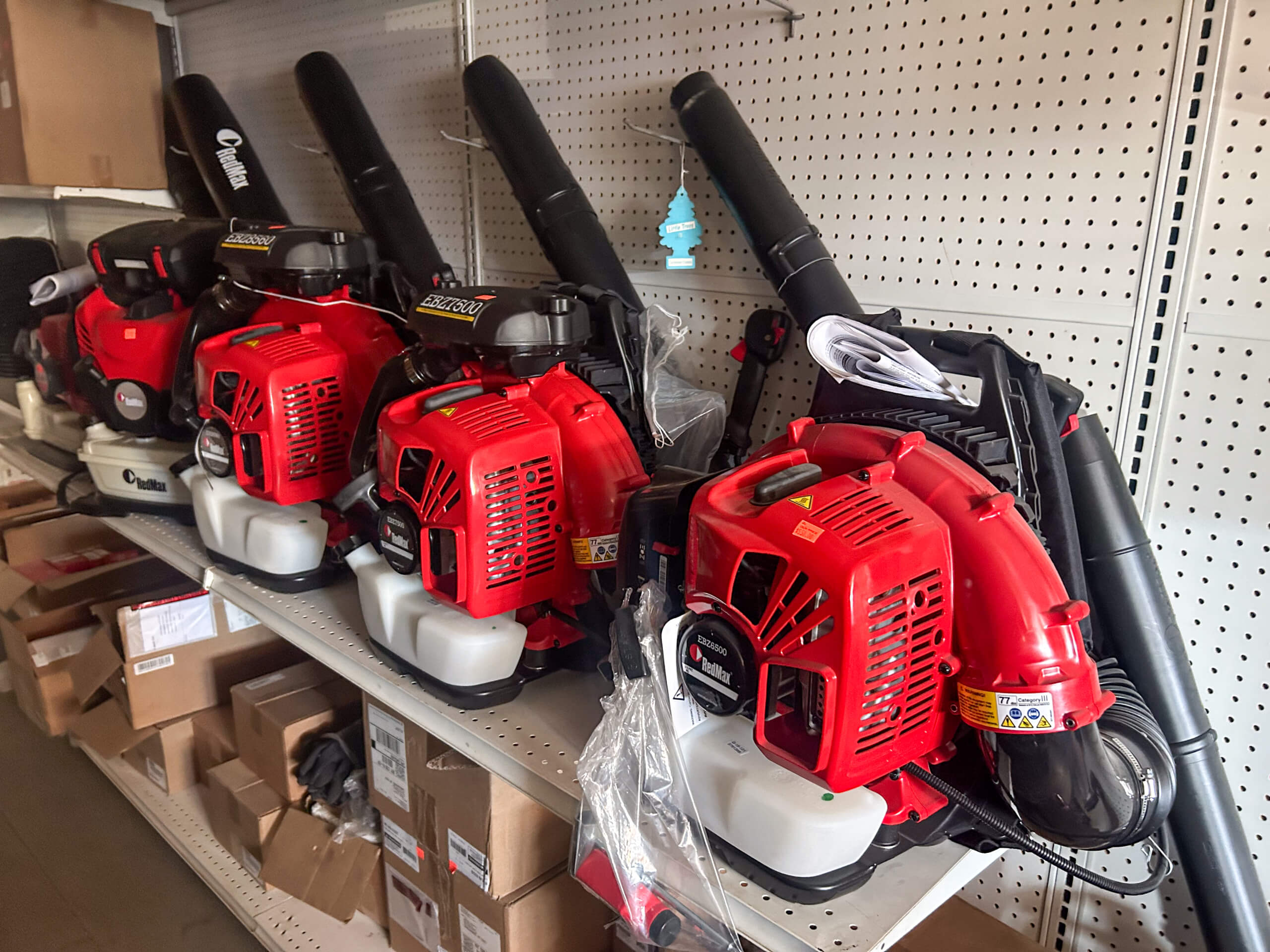 Buel Road Small Engine Repair Canajoharie, NY Four red and black gasoline-powered backpack leaf blowers are lined up on a metal shelf in a store, with boxes stored on the lower shelves.