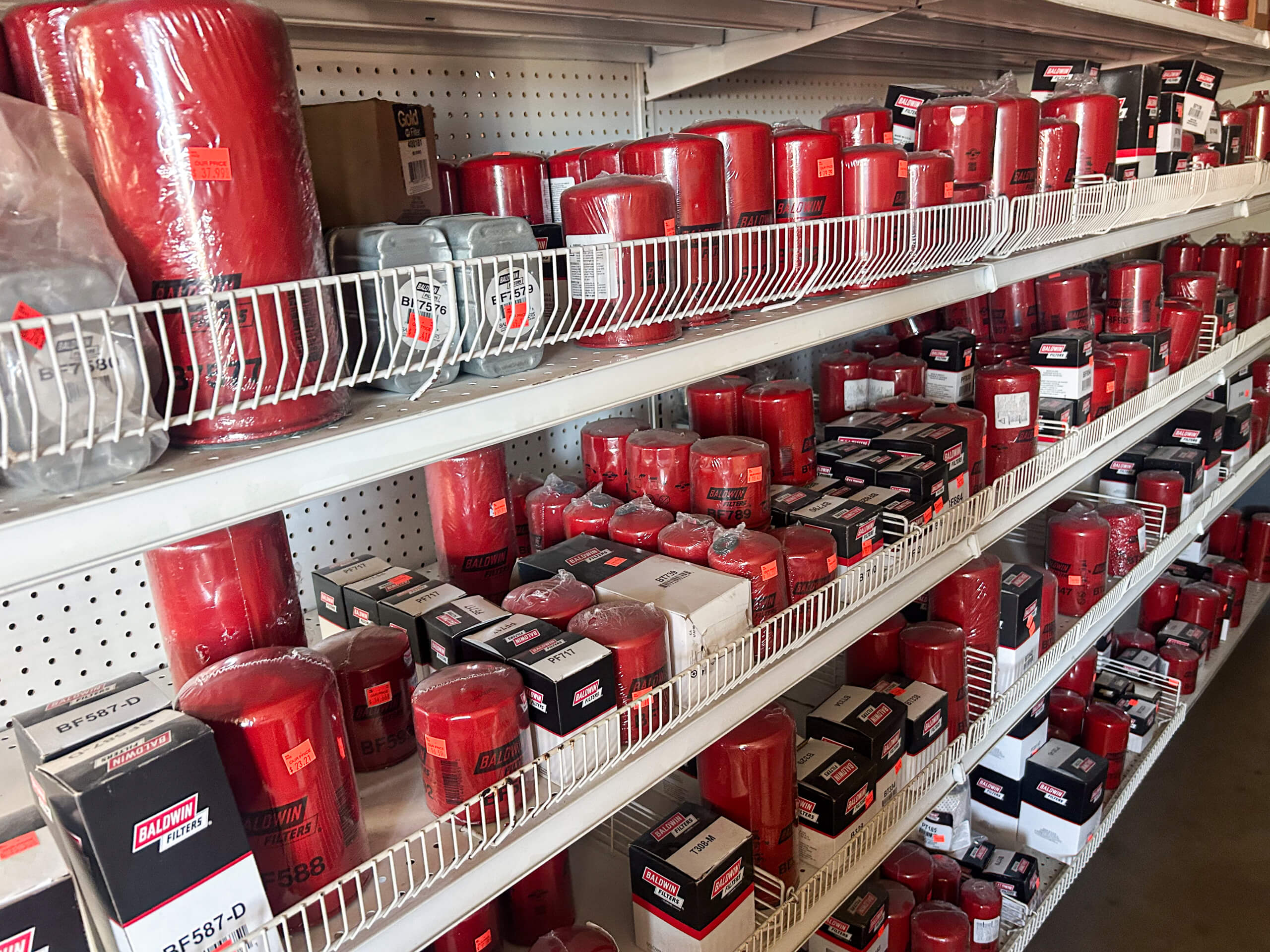 Buel Road Small Engine Repair Canajoharie, NY Shelves stocked with various sizes of red and black Baldwin filters and filter boxes, organized in wire racks in a store aisle.