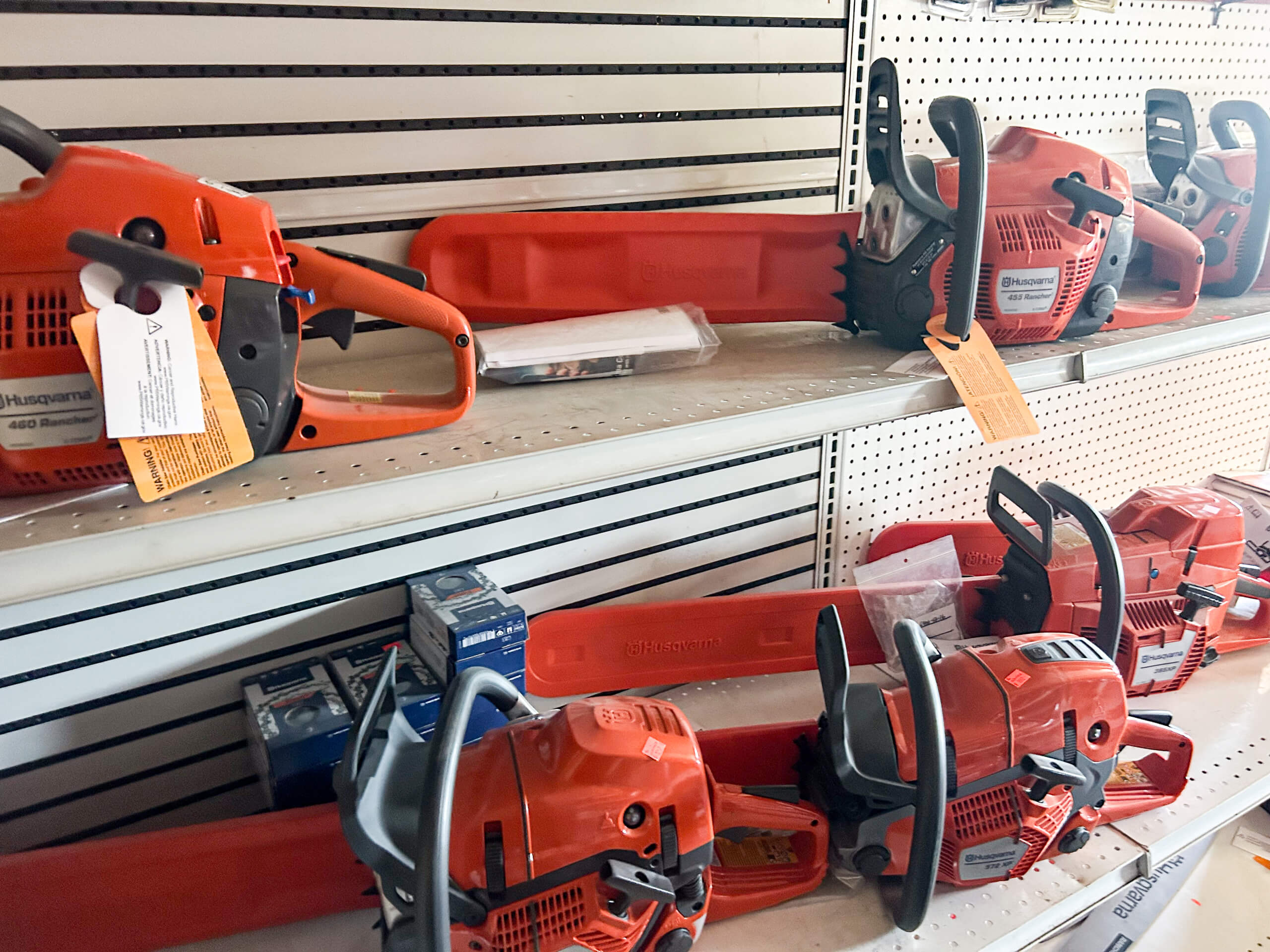 Buel Road Small Engine Repair Canajoharie, NY Several orange chainsaws with protective bar covers are displayed on metal shelves in a hardware store, with product boxes also visible nearby.