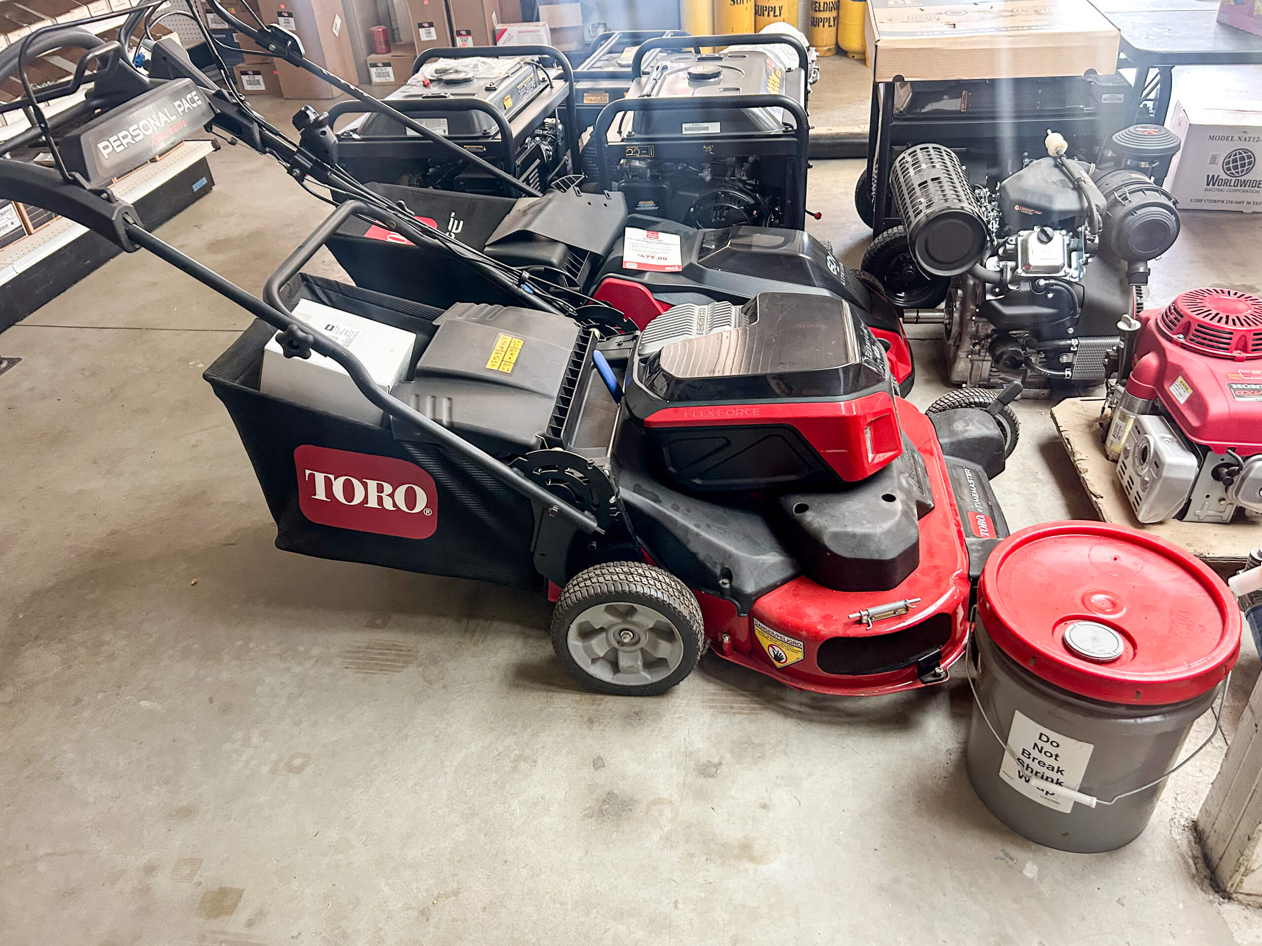 Buel Road Small Engine Repair Canajoharie, NY A red Toro lawn mower is parked indoors next to a red lidded gray bucket and various small engines on a concrete floor.