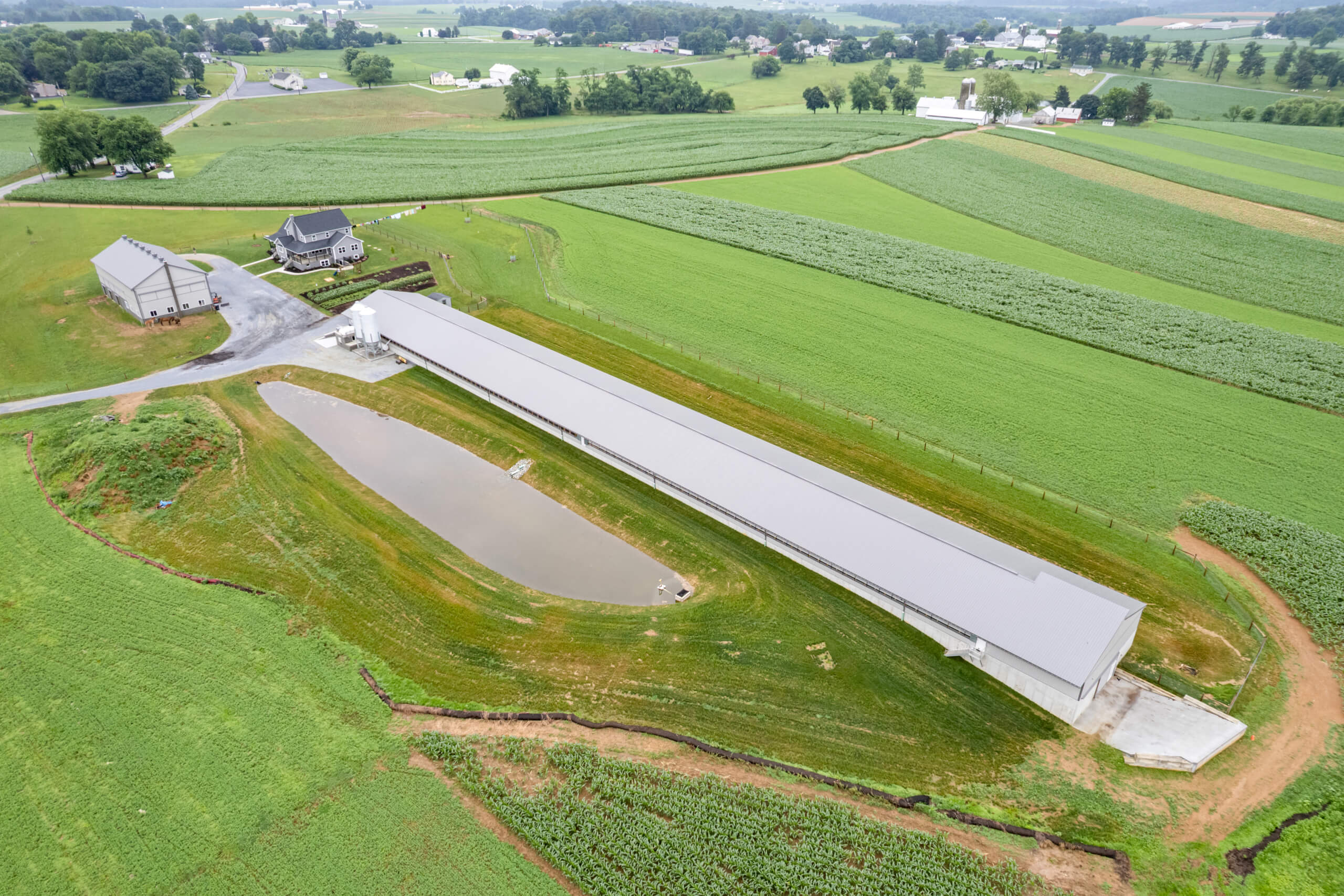 Pine Tree Builders Honeybrook Chester Lancaster County PA Aerial view of a long rectangular agricultural building next to a pond, with a house, a barn, and surrounding green farmland.
