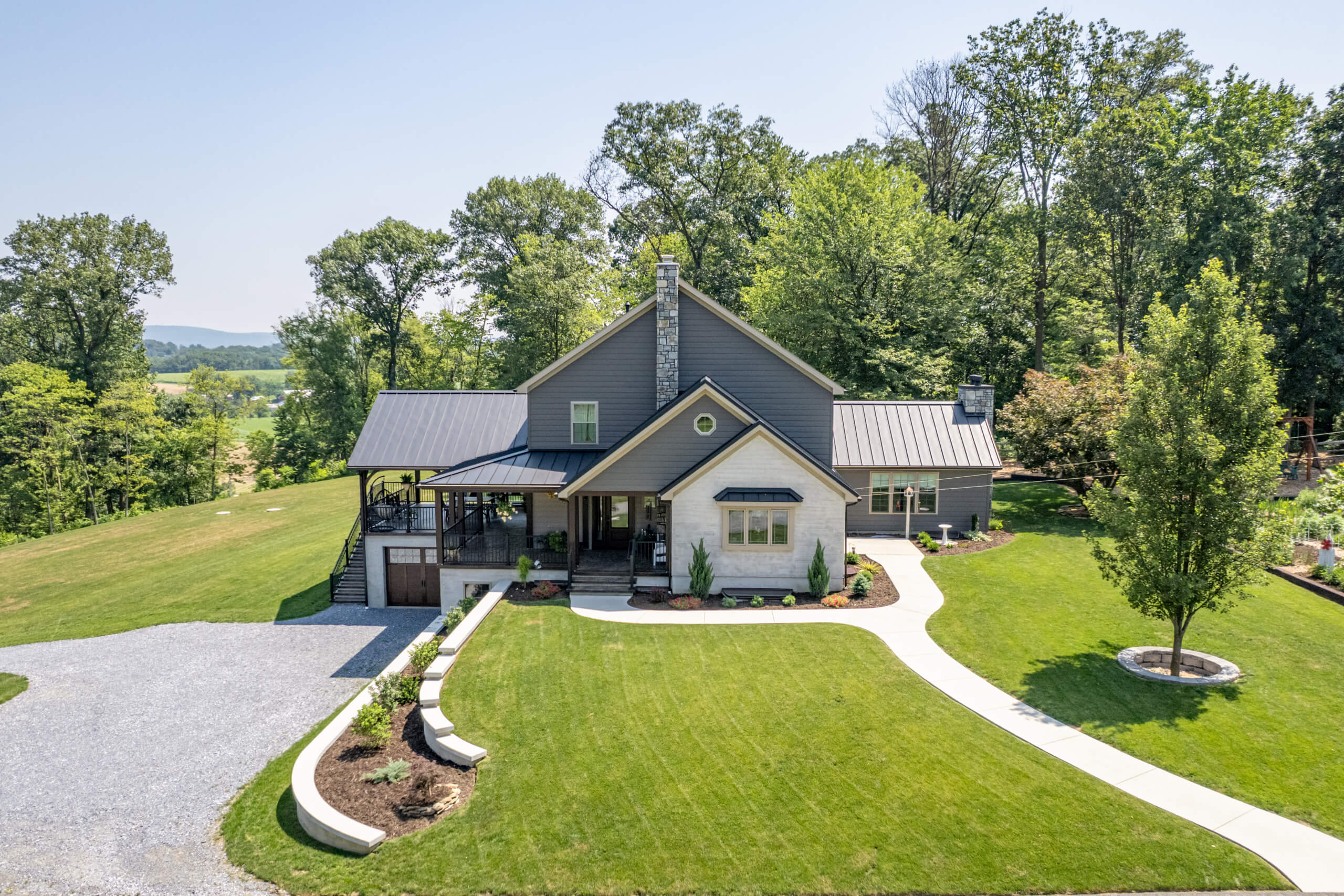 Pine Tree Builders Honeybrook Chester Lancaster County PA A modern farmhouse with gray siding, a metal roof, and covered porch, surrounded by a well-manicured lawn, garden beds, and trees.