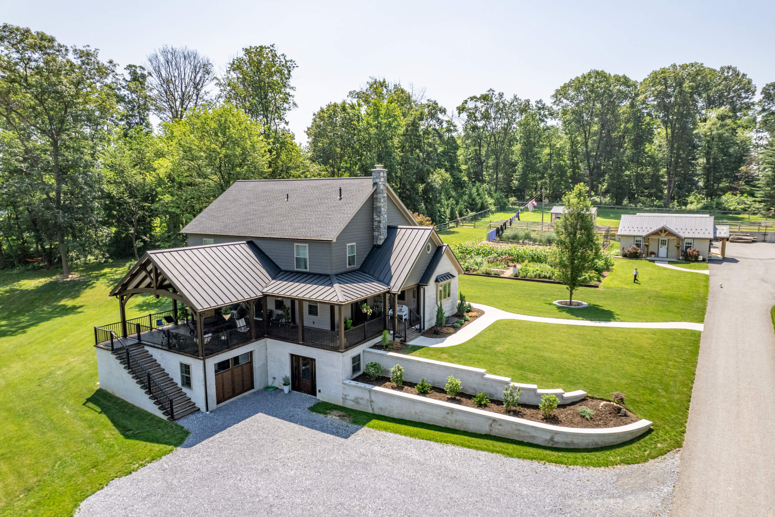 Pine Tree Builders Honeybrook Chester Lancaster County PA Aerial view of a two-story house with a wraparound porch, surrounding lawn, landscaped garden, and a gravel driveway, set in a wooded area with an outbuilding nearby.