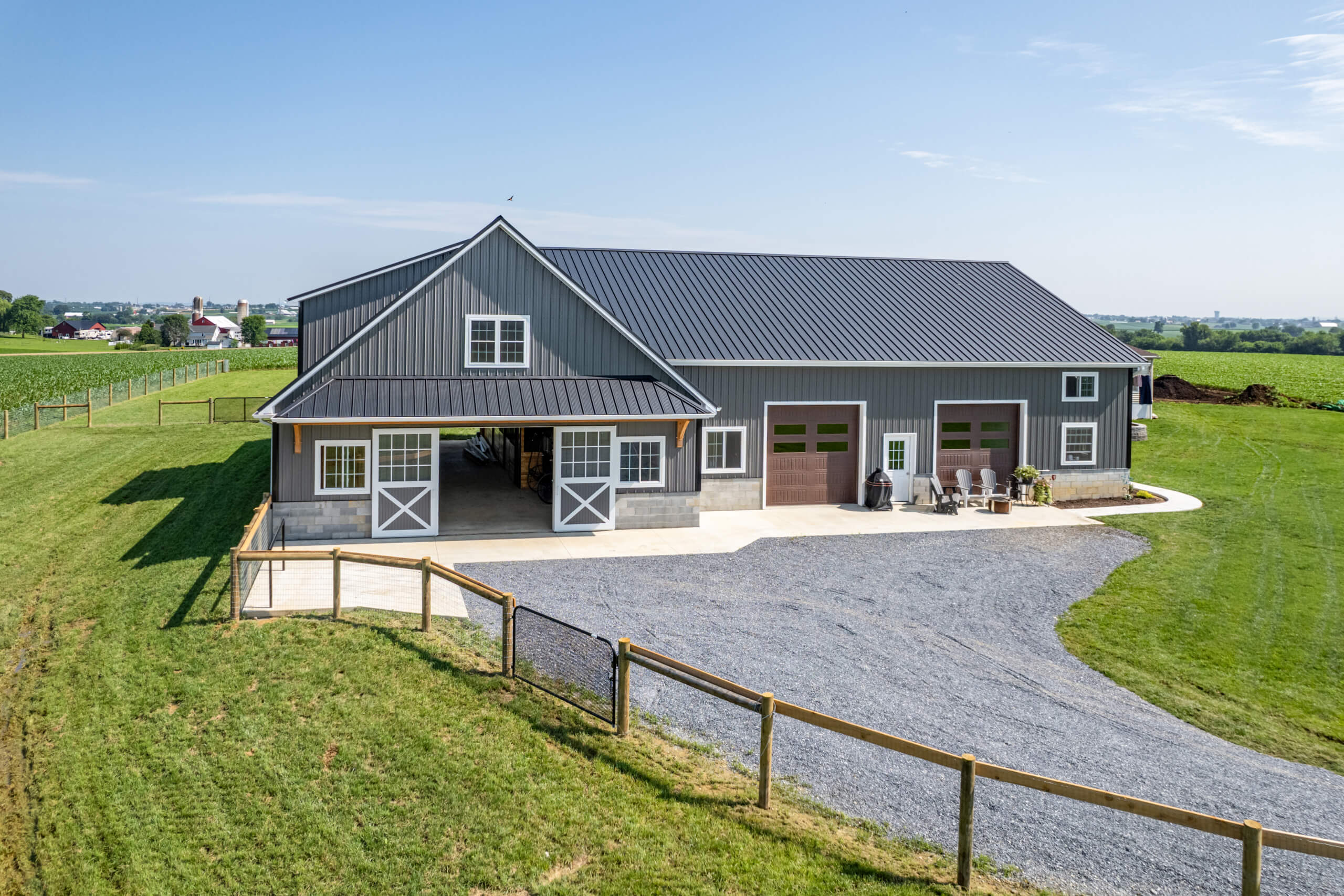 Pine Tree Builders Honeybrook Chester Lancaster County PA A large gray barn with white trim, double garage doors, and gravel driveway sits on a green, fenced-in field under a clear blue sky.