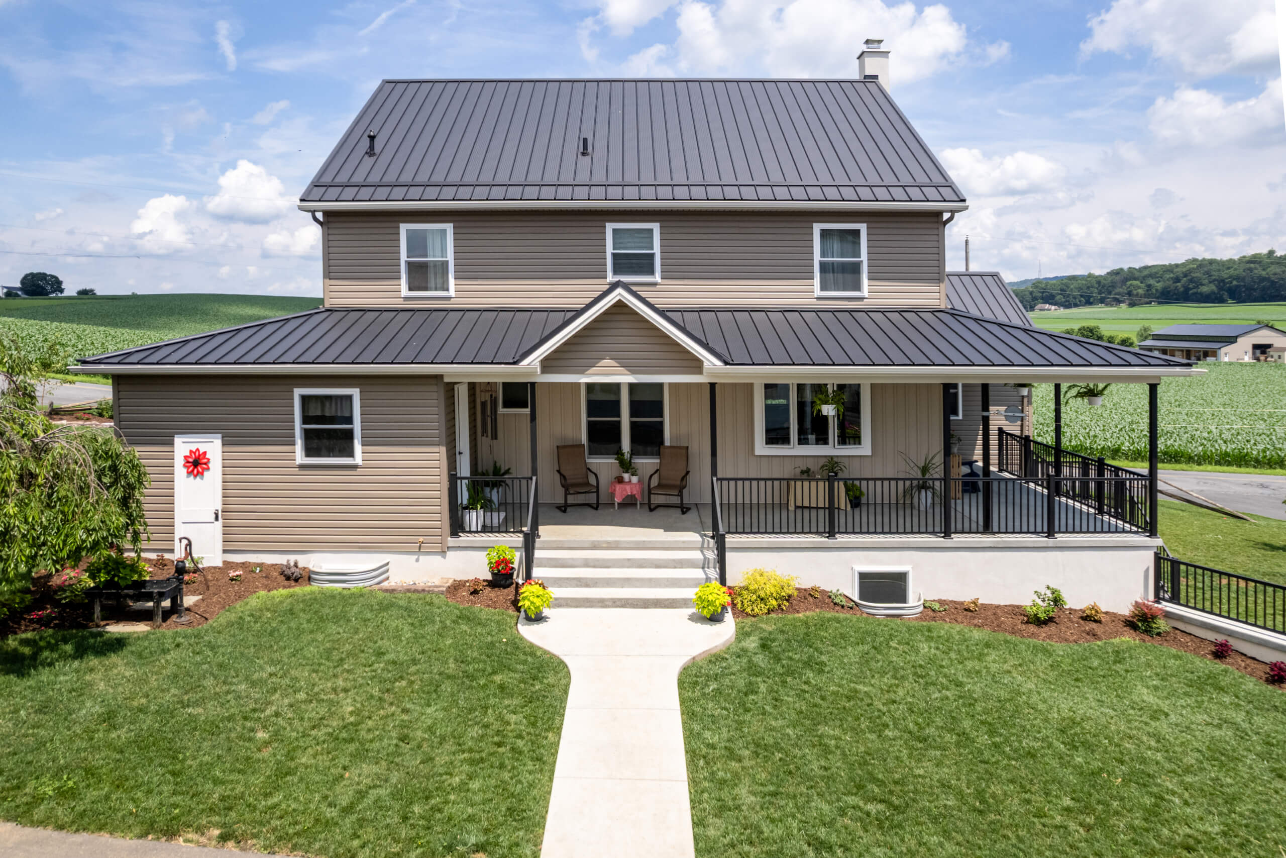 Pine Tree Builders Honeybrook Chester Lancaster County PA Two-story beige house with a dark metal roof, front porch, and steps leading to a walkway, surrounded by green lawn and farmland.