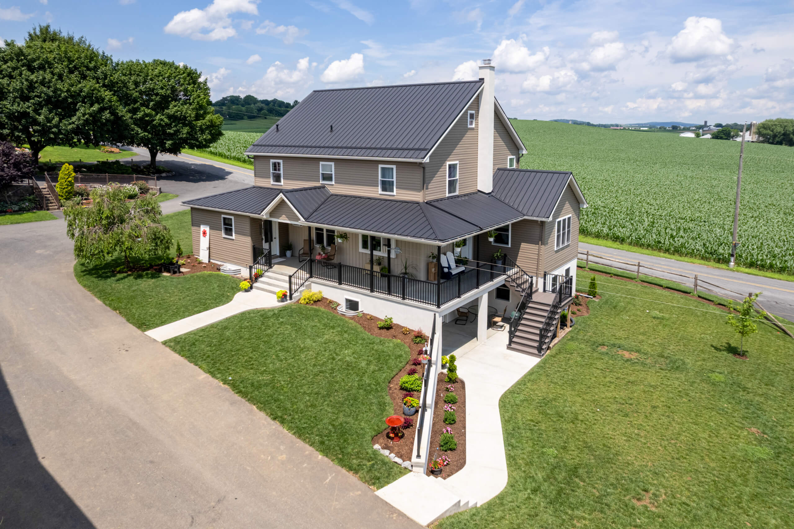 Pine Tree Builders Honeybrook Chester Lancaster County PA Two-story tan house with a black metal roof, large wraparound porch, landscaped yard, and surrounded by fields under a partly cloudy sky.