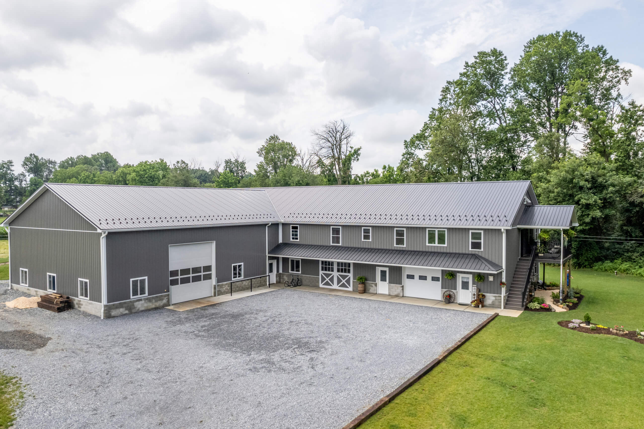 Pine Tree Builders Honeybrook Chester Lancaster County PA Large gray metal barn with multiple garage doors, gravel driveway, green lawn, and trees in the background under a cloudy sky.