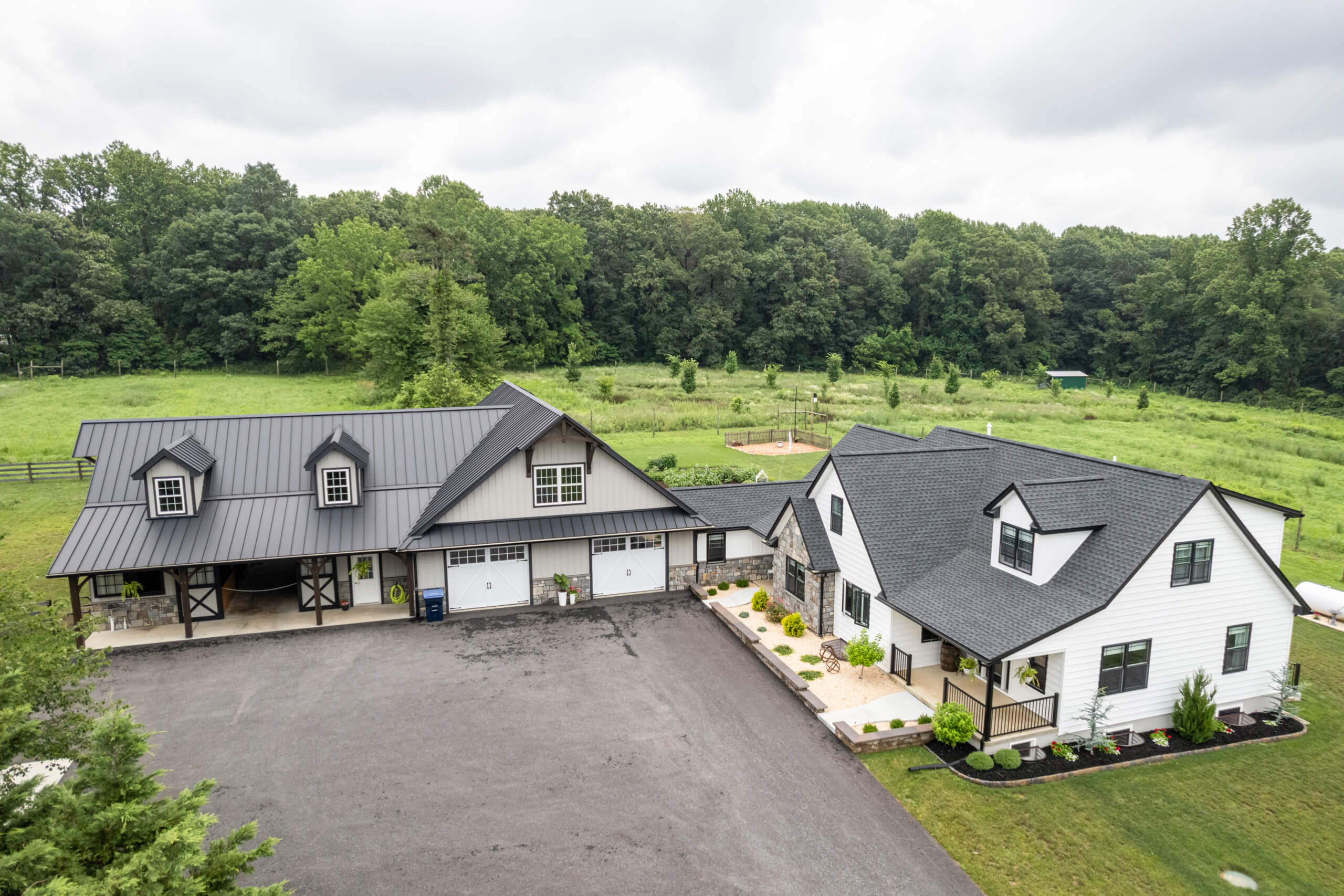 Pine Tree Builders Honeybrook Chester Lancaster County PA Aerial view of a modern farmhouse with a white exterior, black roof, large driveway, attached garage, and surrounding greenery.