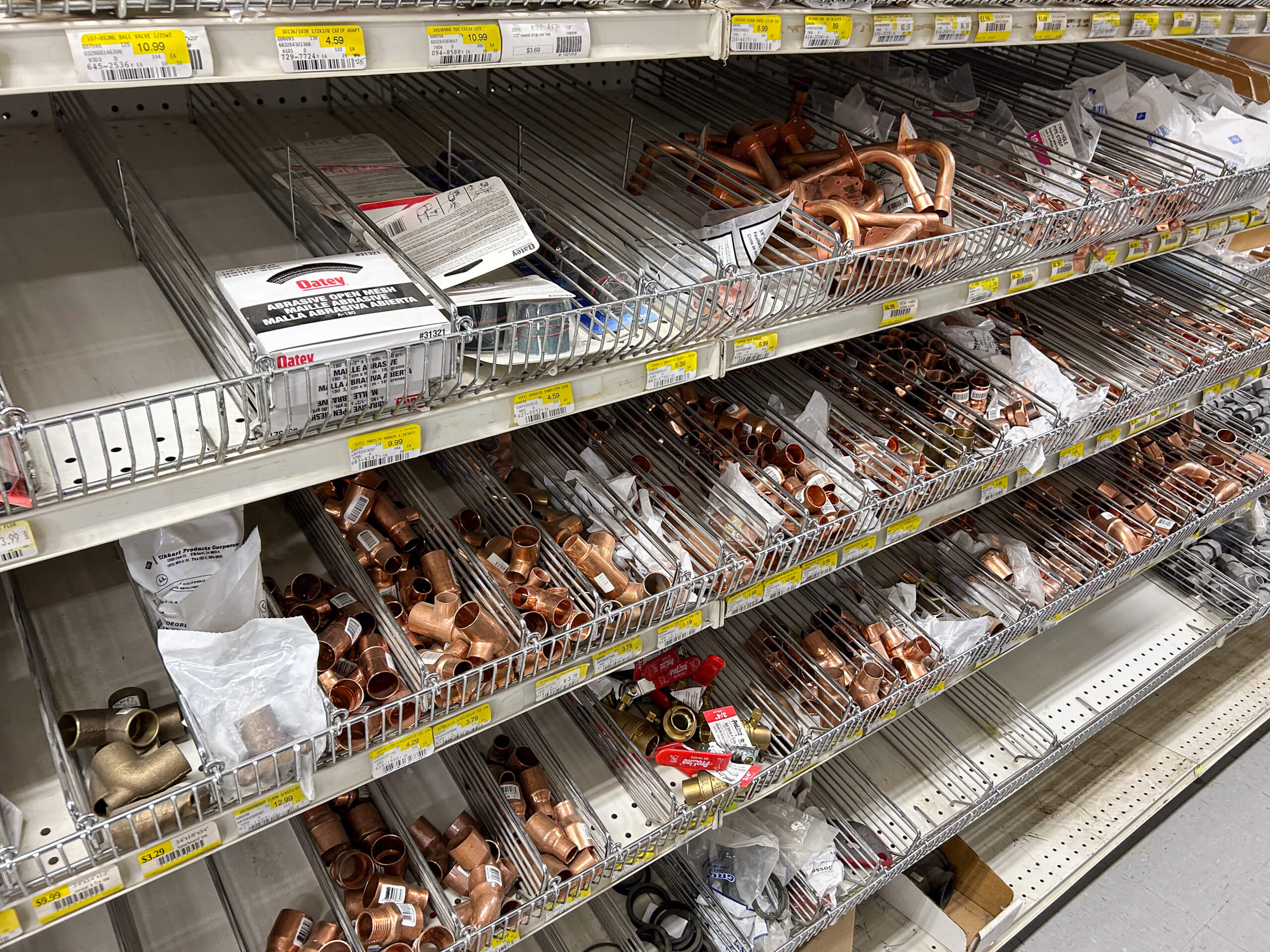 Benny's Hardware Barnett MO Metal plumbing fittings, valves, and connectors are displayed in partially stocked wire bins on shelves in a hardware store aisle.