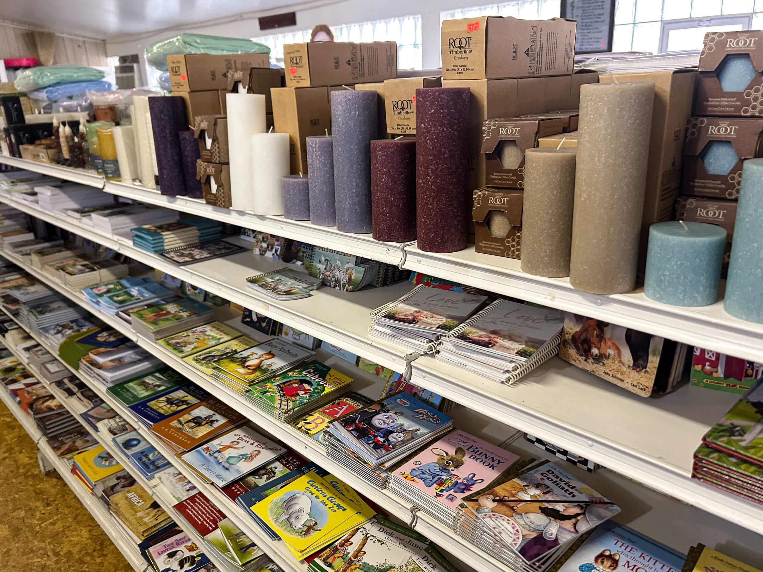Miller's Country Store Grabill IN Shelves in a store display pillar candles, boxes, stacks of books, and magazines arranged neatly in rows.