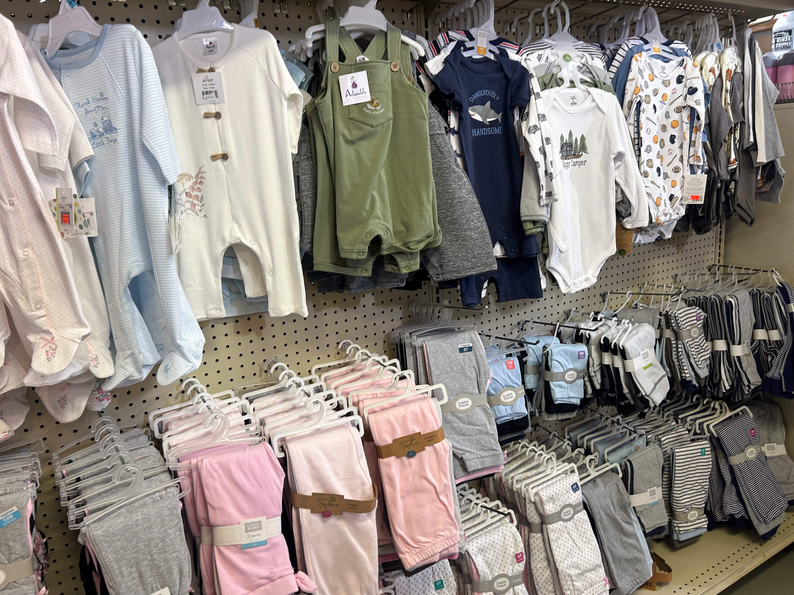 Miller's Country Store Grabill IN Shelves and racks display assorted baby clothes, including onesies, rompers, pants, and socks in various colors and patterns in a retail store.