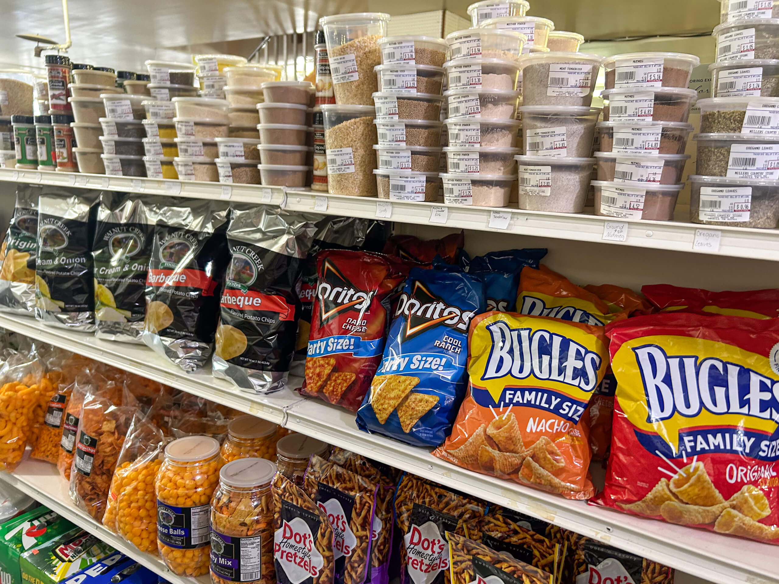 Miller's Country Store Grabill IN Shelves stocked with assorted chips, pretzels, cheese balls, and clear containers of bulk spices or seasonings in a grocery store aisle.