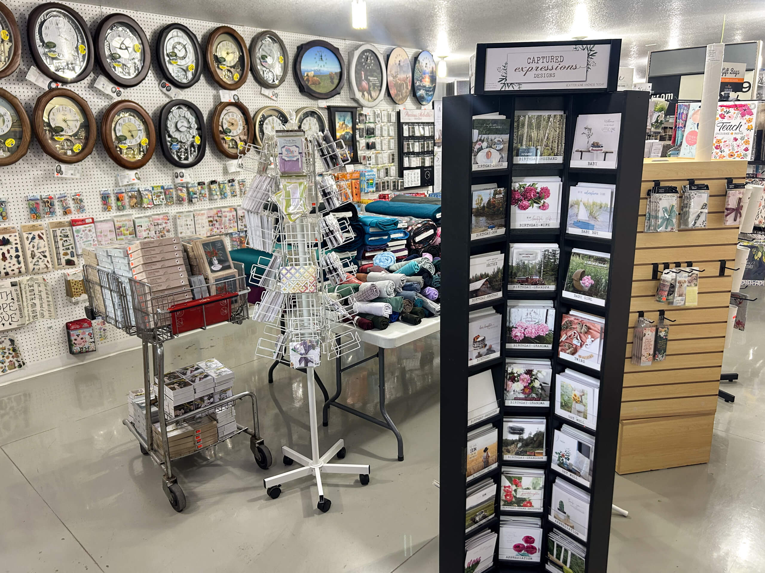 Miller's Country Store Grabill IN A store interior displays clocks on the wall, greeting cards on a rack, folded blankets, puzzles, and various gift items on tables and shelves.