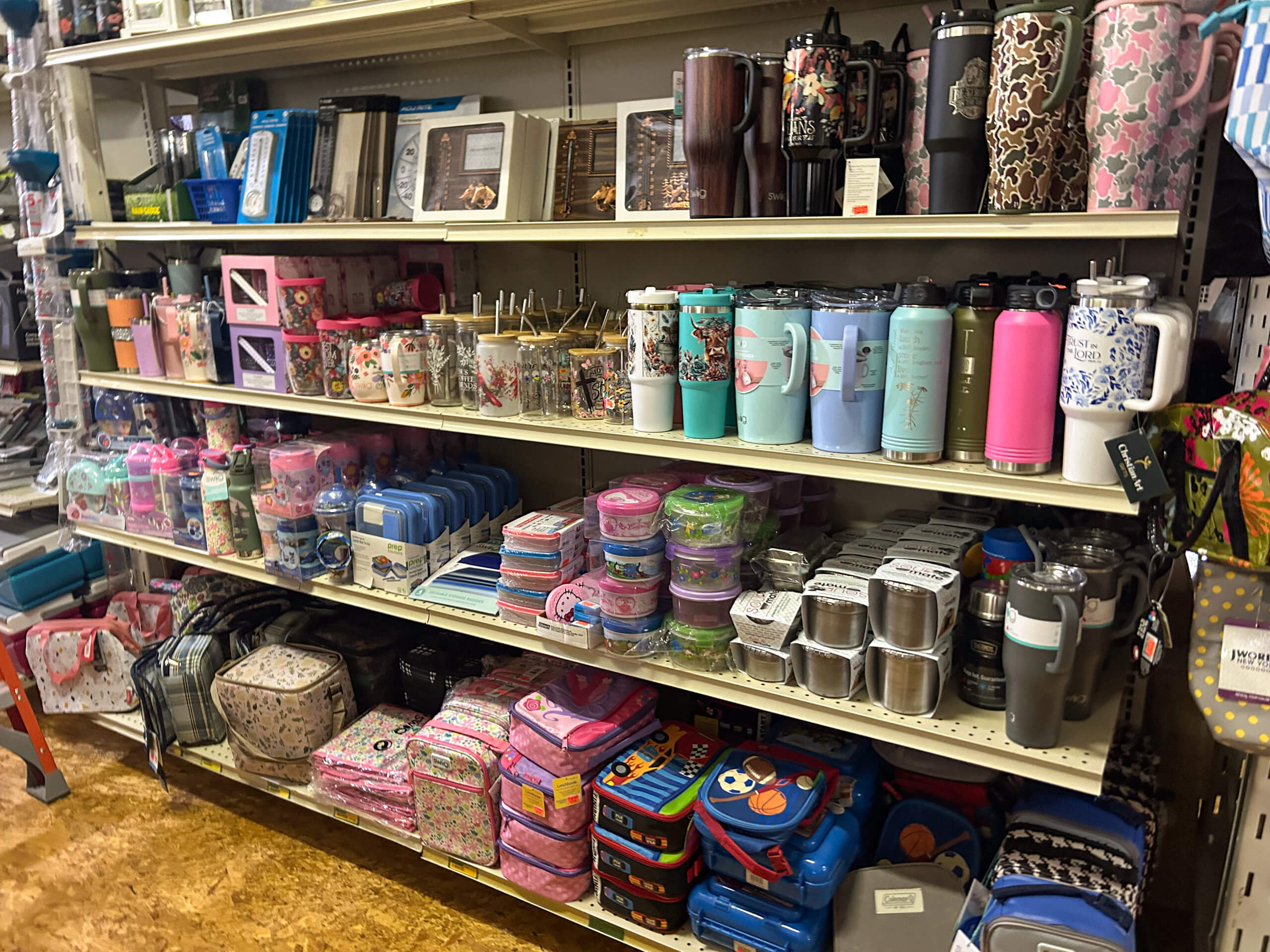 Miller's Country Store Grabill IN Store shelves display an assortment of lunch boxes, food containers, travel mugs, water bottles, thermoses, and insulated bags in various colors and patterns.