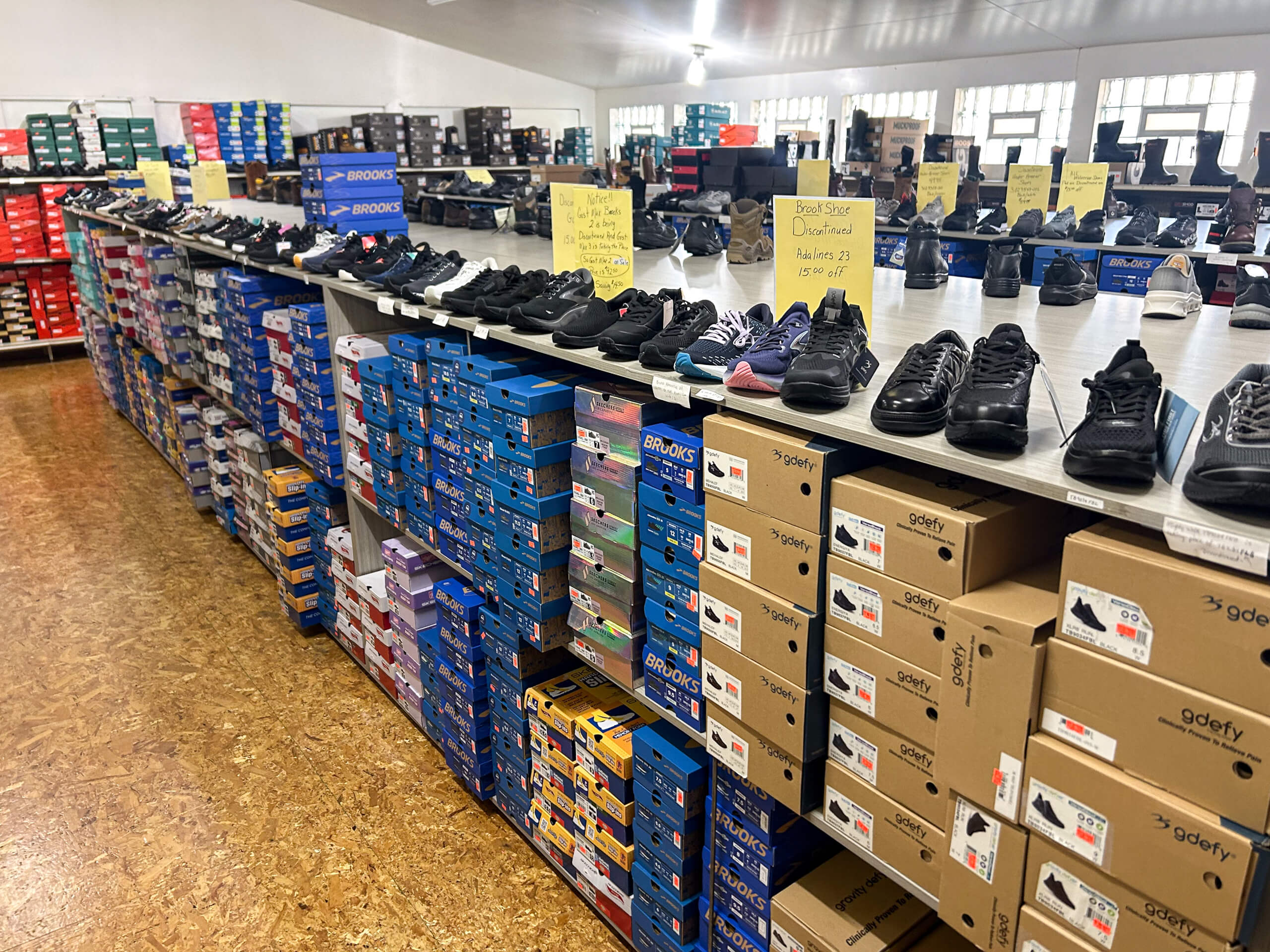 Miller's Country Store Grabill IN Rows of athletic shoes displayed on top of stacked shoeboxes in a brightly lit shoe store; yellow sale signs are visible on the table.