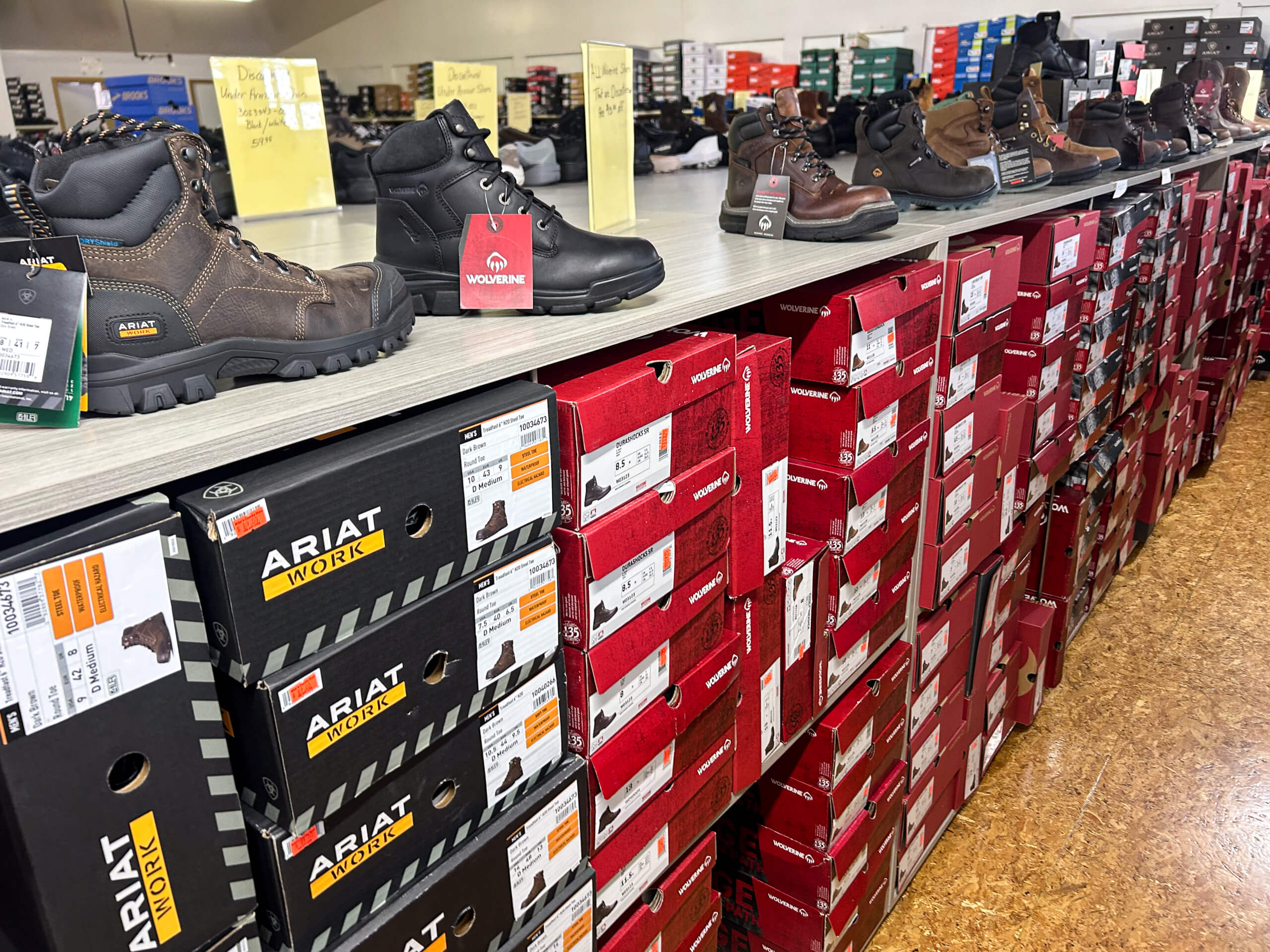Miller's Country Store Grabill IN Assortment of work boots displayed on a store shelf above stacks of boxed Ariat brand footwear in a retail setting.